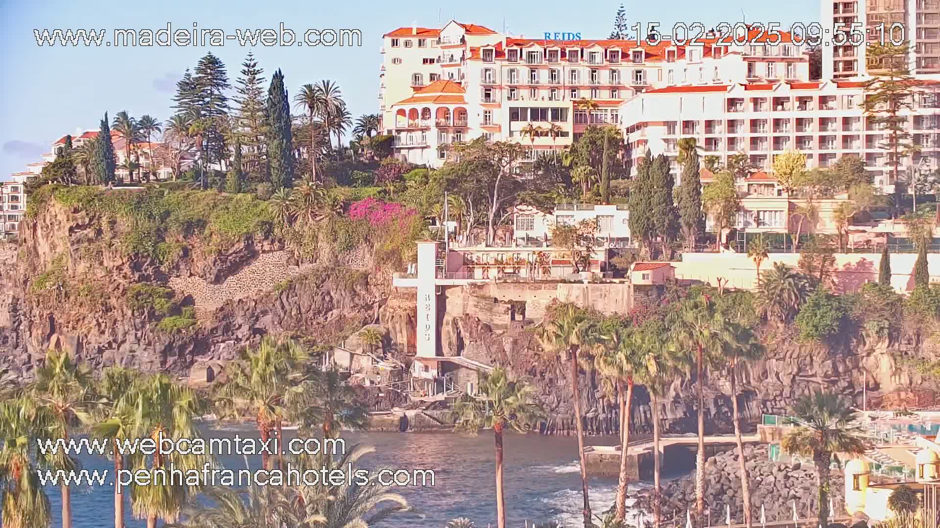 A large hotel sits atop a rocky cliff overlooking a calm body of water, surrounded by lush greenery and palm trees under a sunny sky.