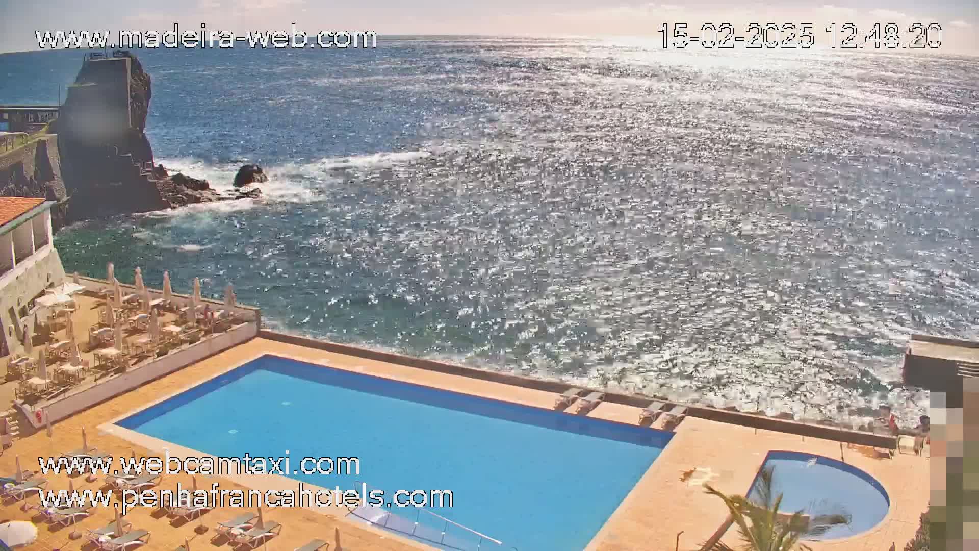 A hotel pool area with a patio and outdoor seating overlooks a rocky coast and a bright, sparkling ocean under sunny skies.