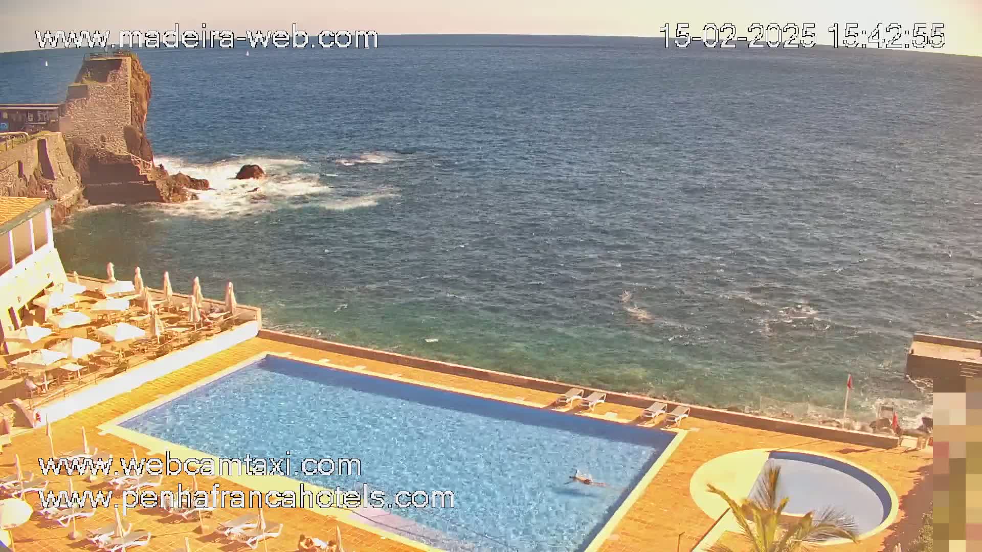 An outdoor swimming pool and a smaller circular pool are situated on a hotel patio overlooking a calm ocean and a rocky coastline on a sunny day.