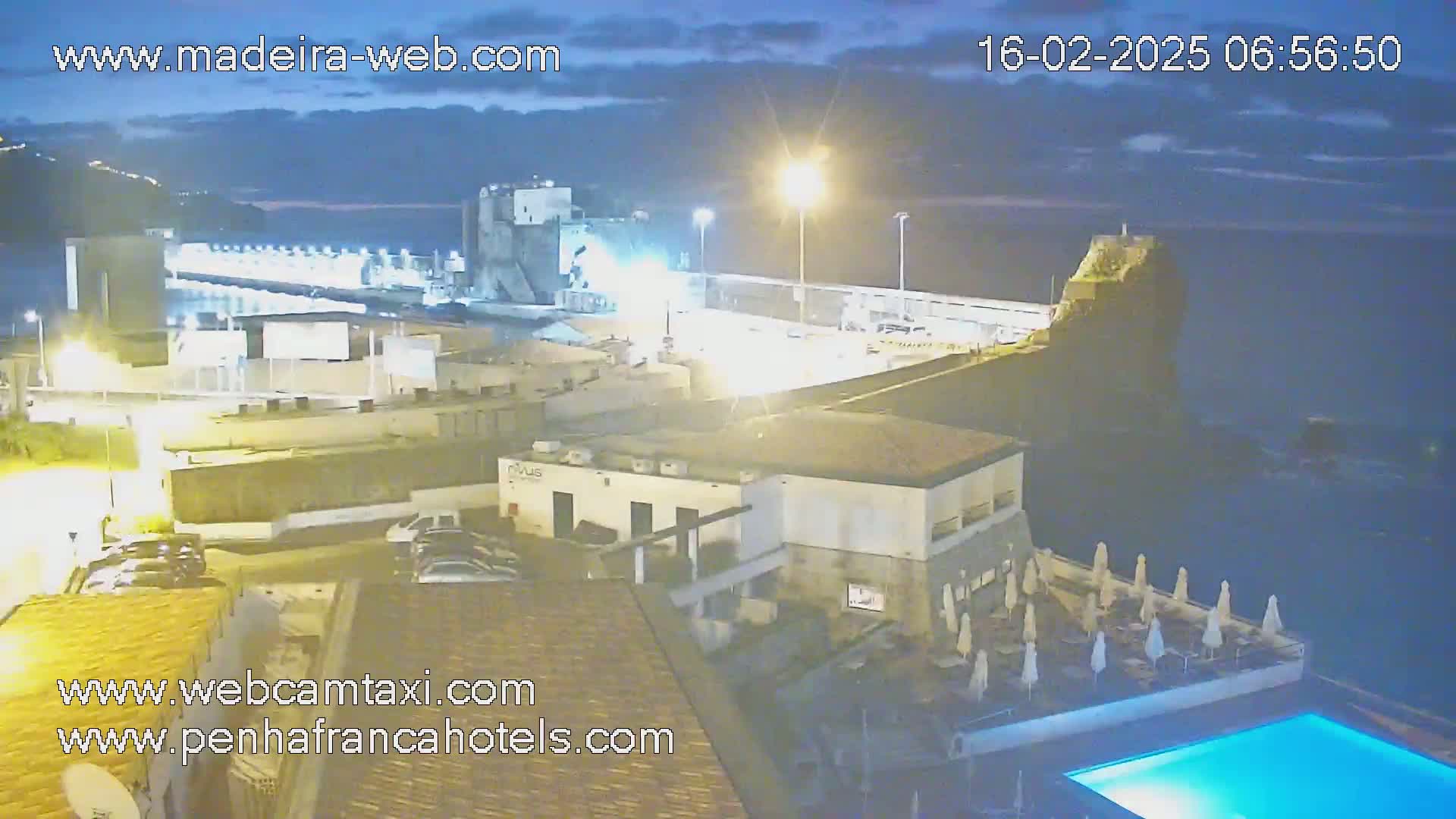 An outdoor nighttime scene shows a coastal area with buildings, a harbor, a pool, and calm water under a partly cloudy sky.