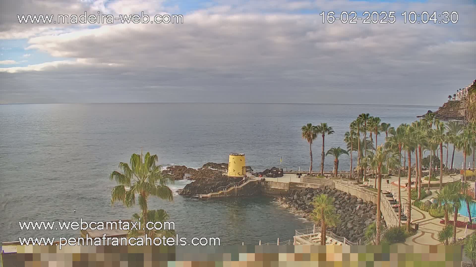 A mostly cloudy day reveals a coastal scene with palm trees, a rocky shoreline, and calm ocean water.