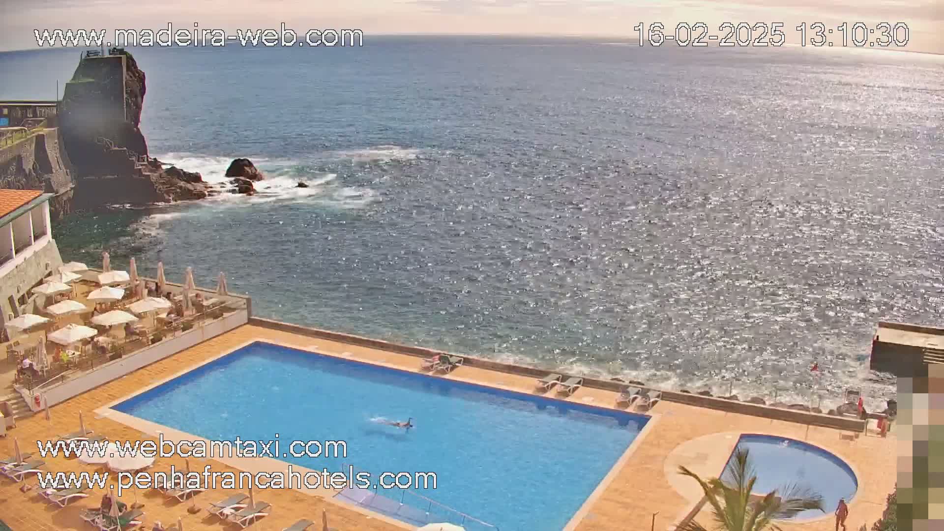 A hotel pool and patio area overlooks a rocky coastline and a sparkling ocean under a sunny sky.