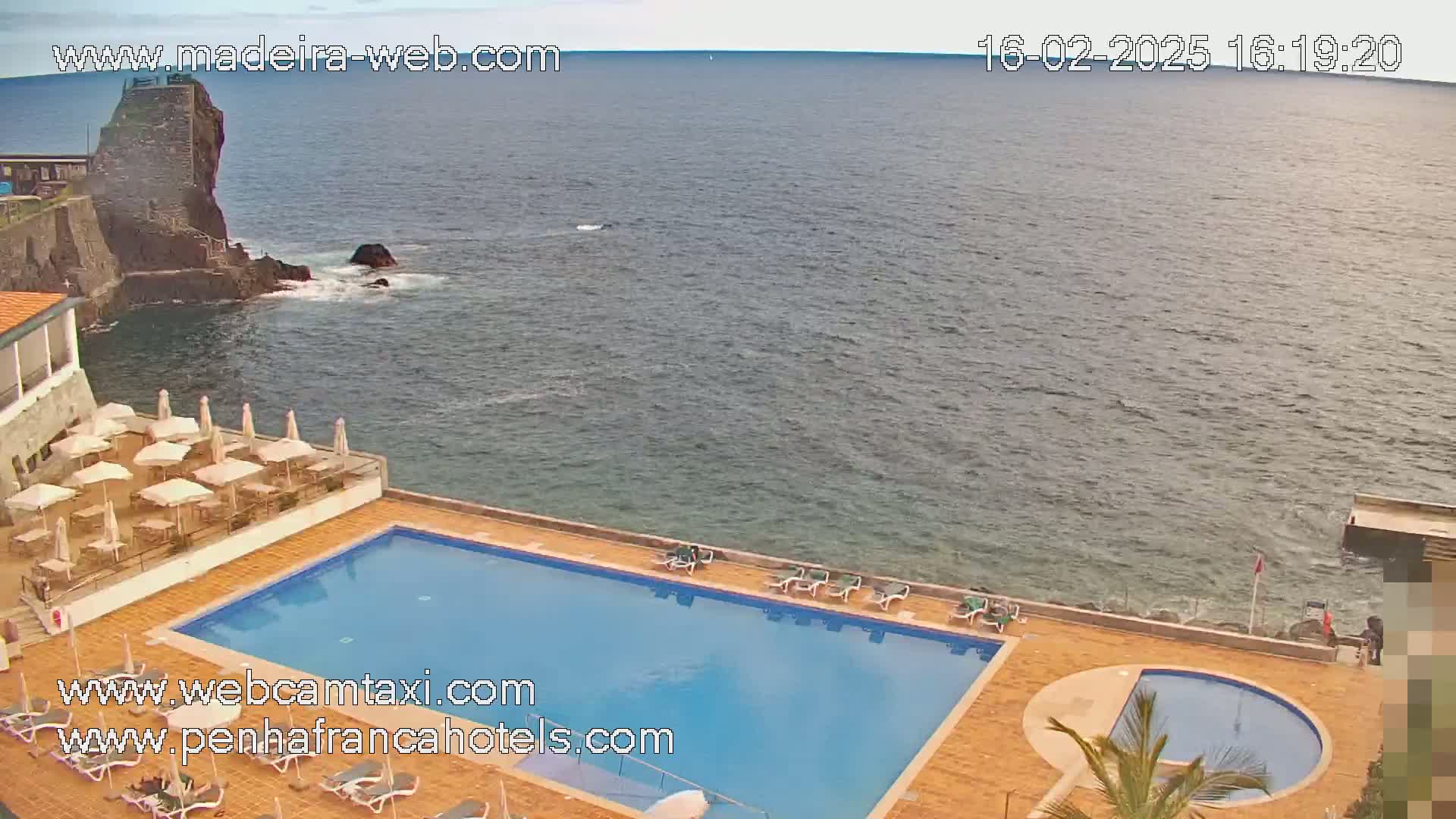 An outdoor swimming pool and a smaller round pool are situated on a seaside resort next to dark-grey rocks and a calm ocean under an overcast sky.