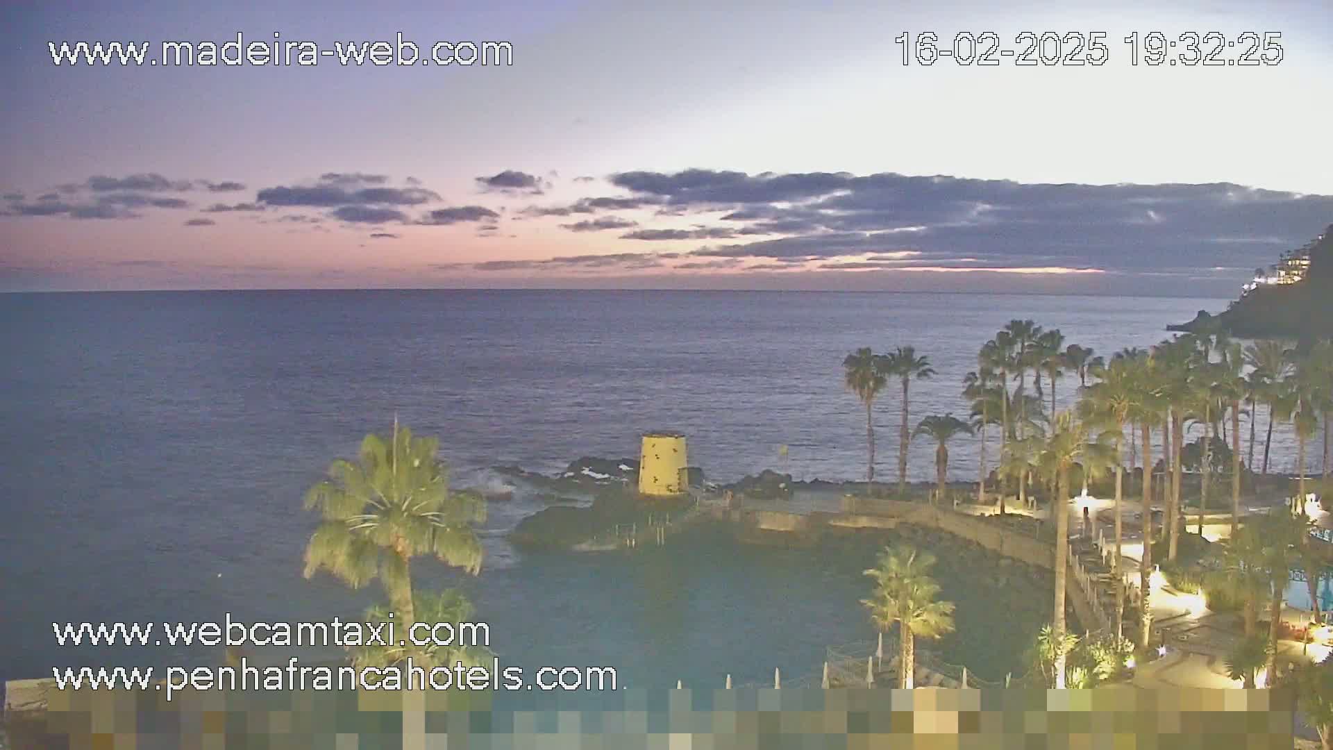 A twilight coastal scene shows a calm ocean under a partly cloudy sky, with palm trees lining a rocky shoreline and a small yellow structure near the water's edge.