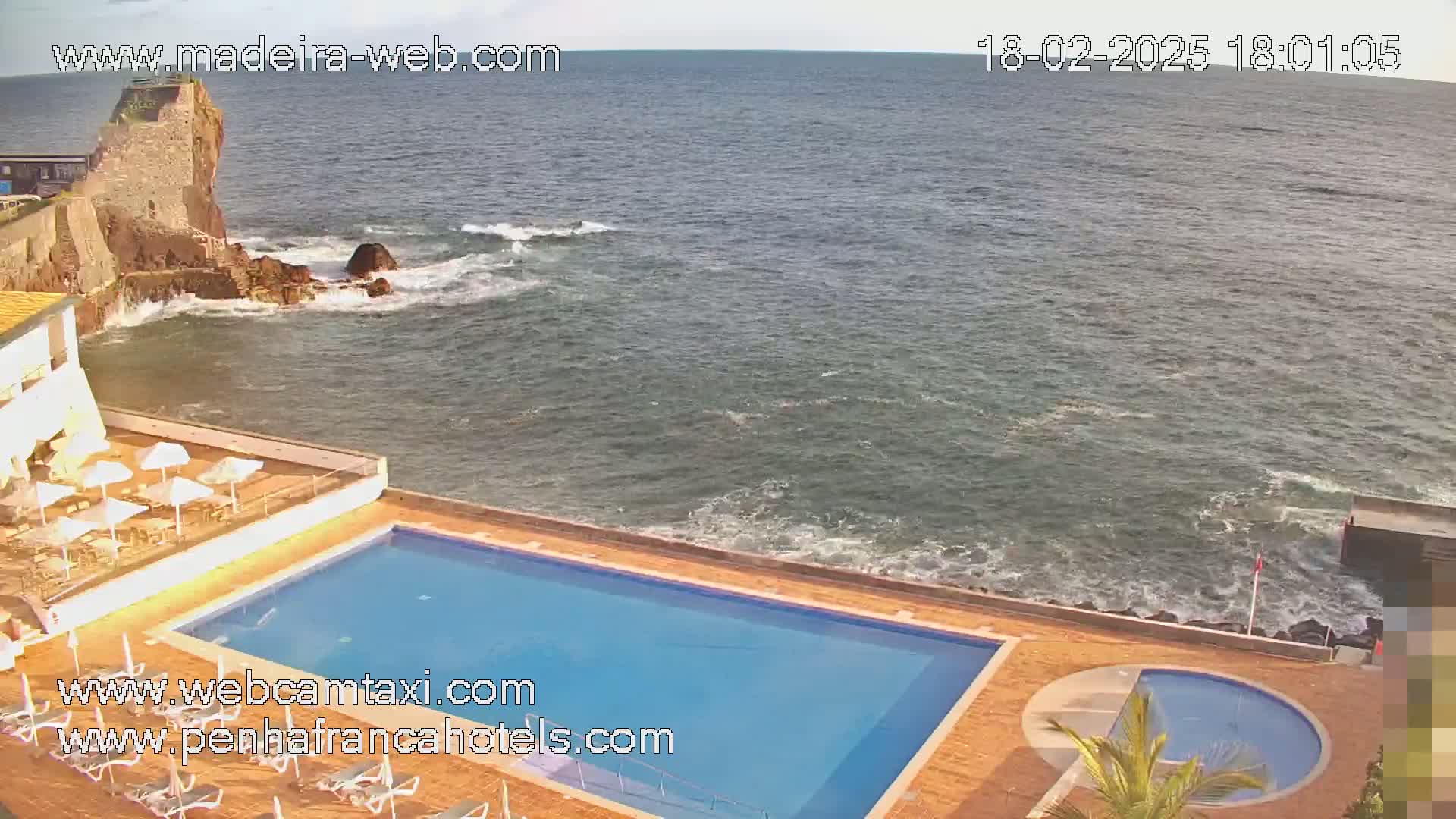 An ocean view overlooking a resort pool and a rocky coastline under a mostly clear sky.
