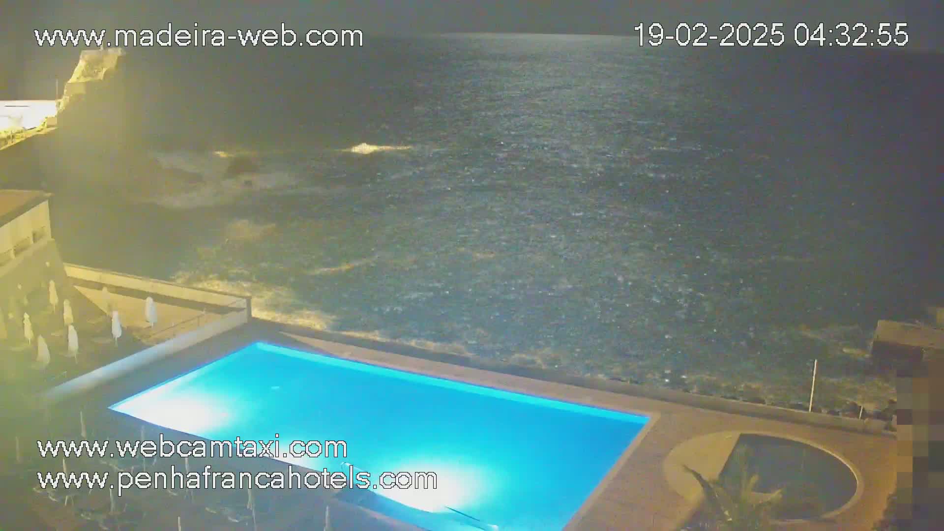 At night, a lit pool sits next to a dark ocean with waves crashing against a rocky shore.