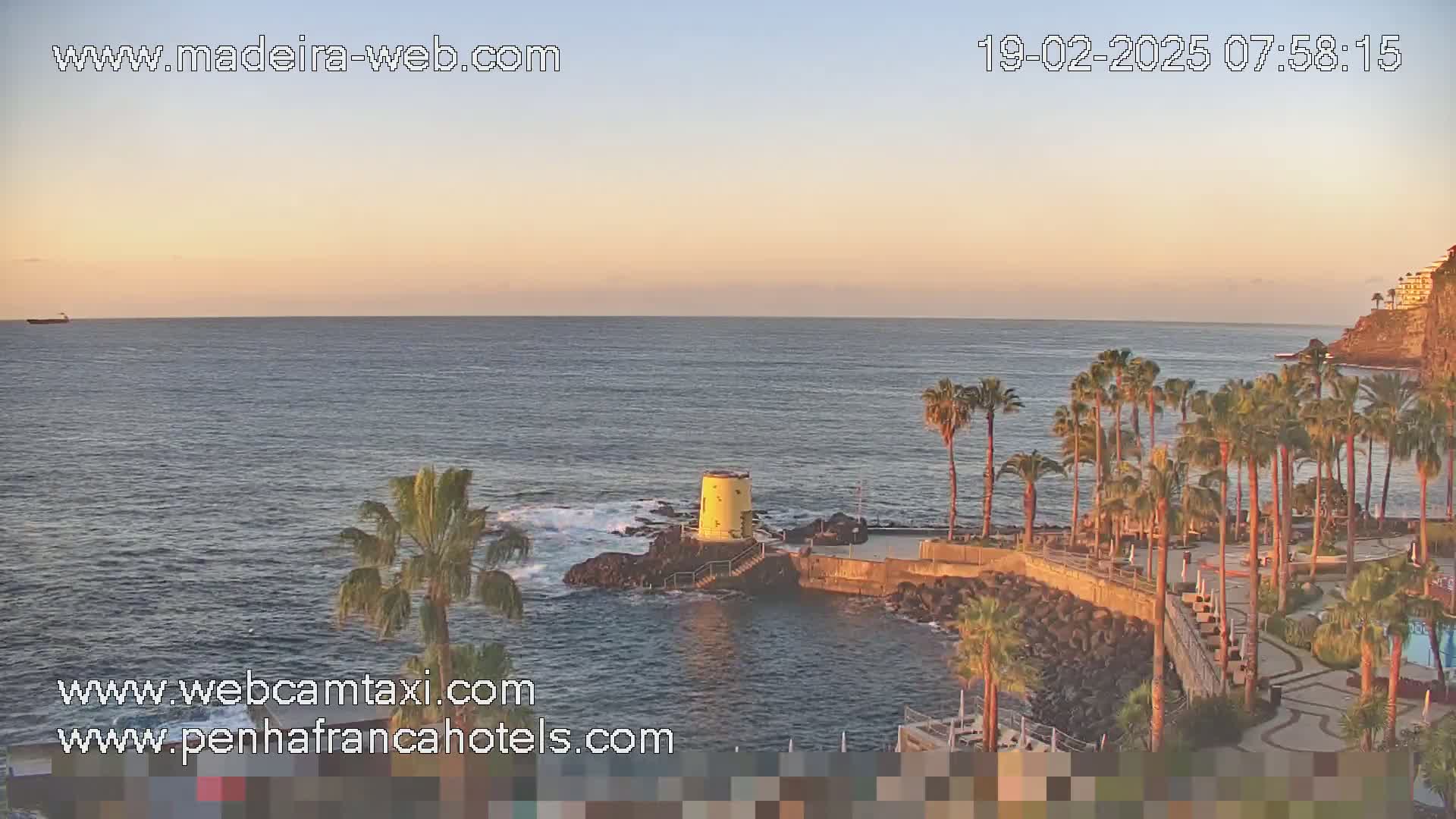 A calm ocean at sunrise is viewed from a seaside location with palm trees, a small rocky outcropping, and a yellow structure.
