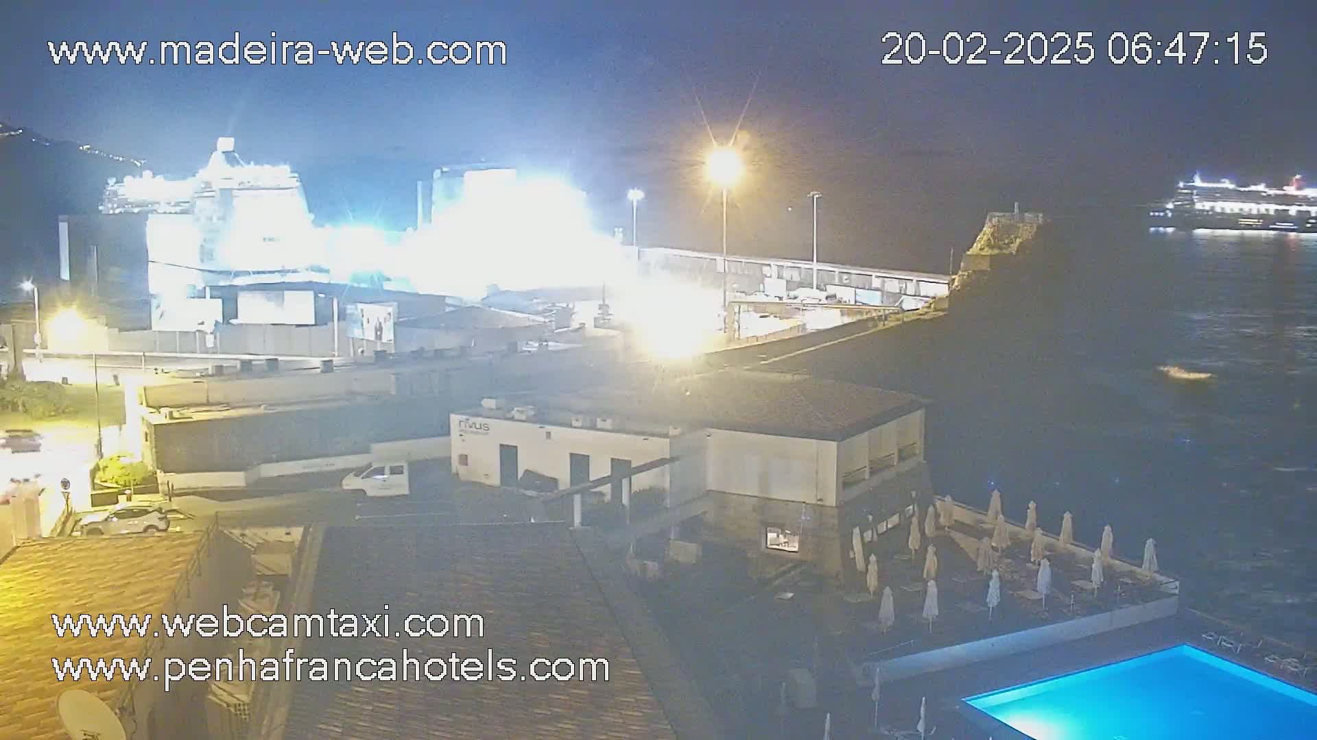 A nighttime harbor scene shows a large cruise ship docked near a smaller one in the water, with brightly lit buildings, a pool, and closed umbrellas on a patio.
