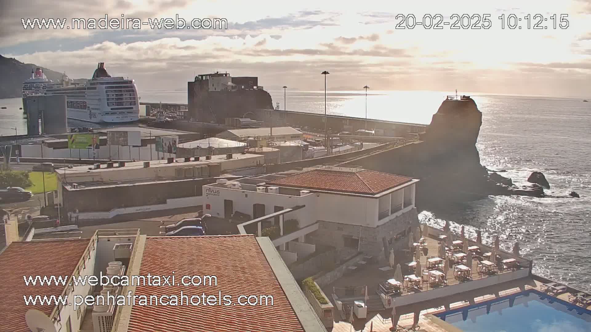 A cruise ship is docked near a coastal town under partly cloudy skies, with a rocky outcrop and ocean visible in the background.