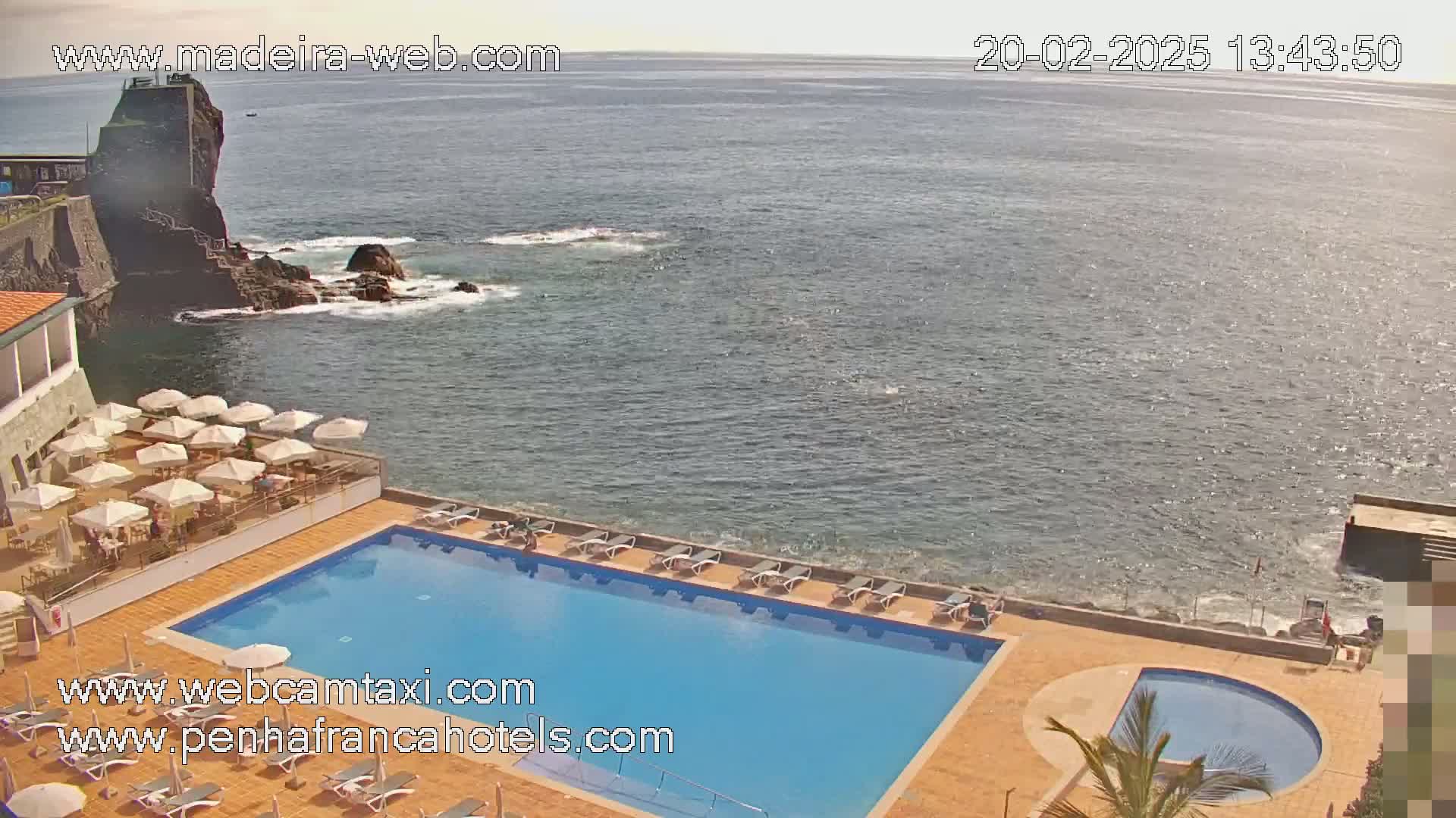 A resort pool area with lounge chairs and umbrellas overlooks a calm ocean next to a rocky coastline on a sunny day.