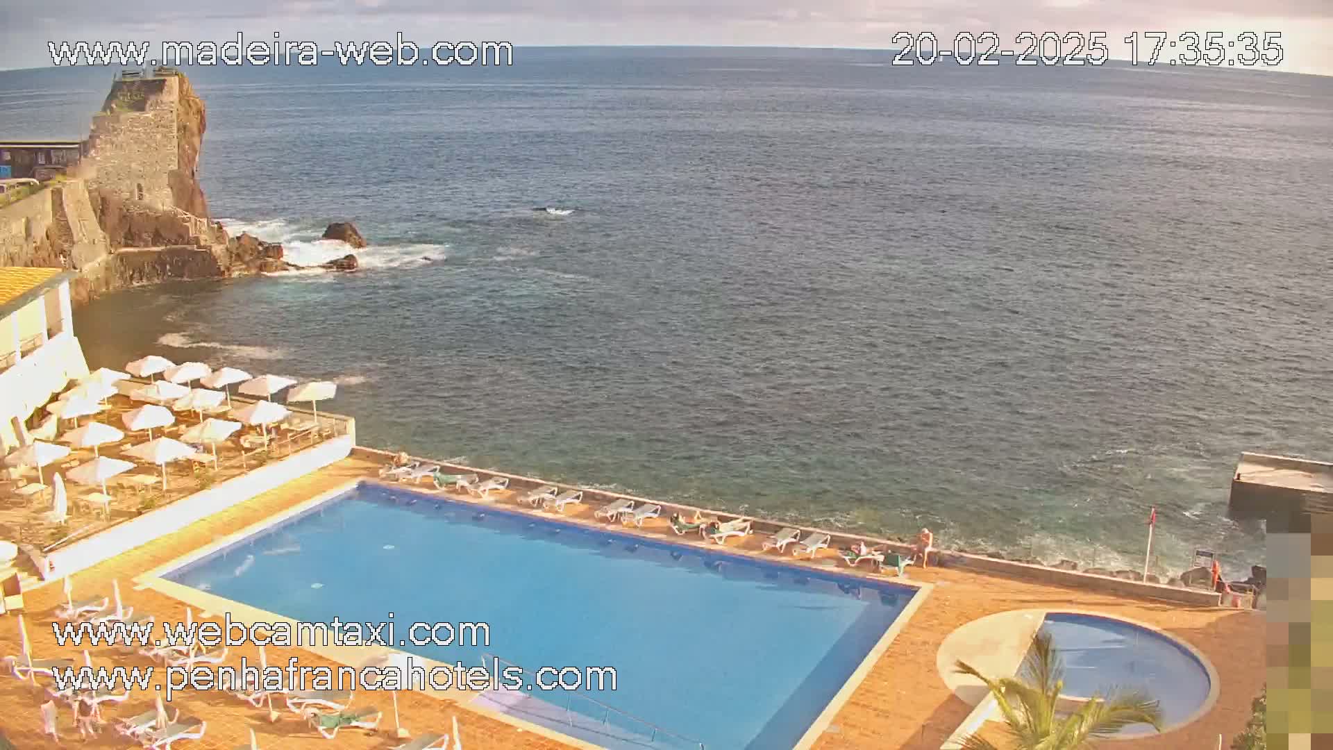 An outdoor swimming pool and a smaller adjacent pool are situated on a hotel overlooking a calm ocean next to a rocky coastline under a mostly clear sky.