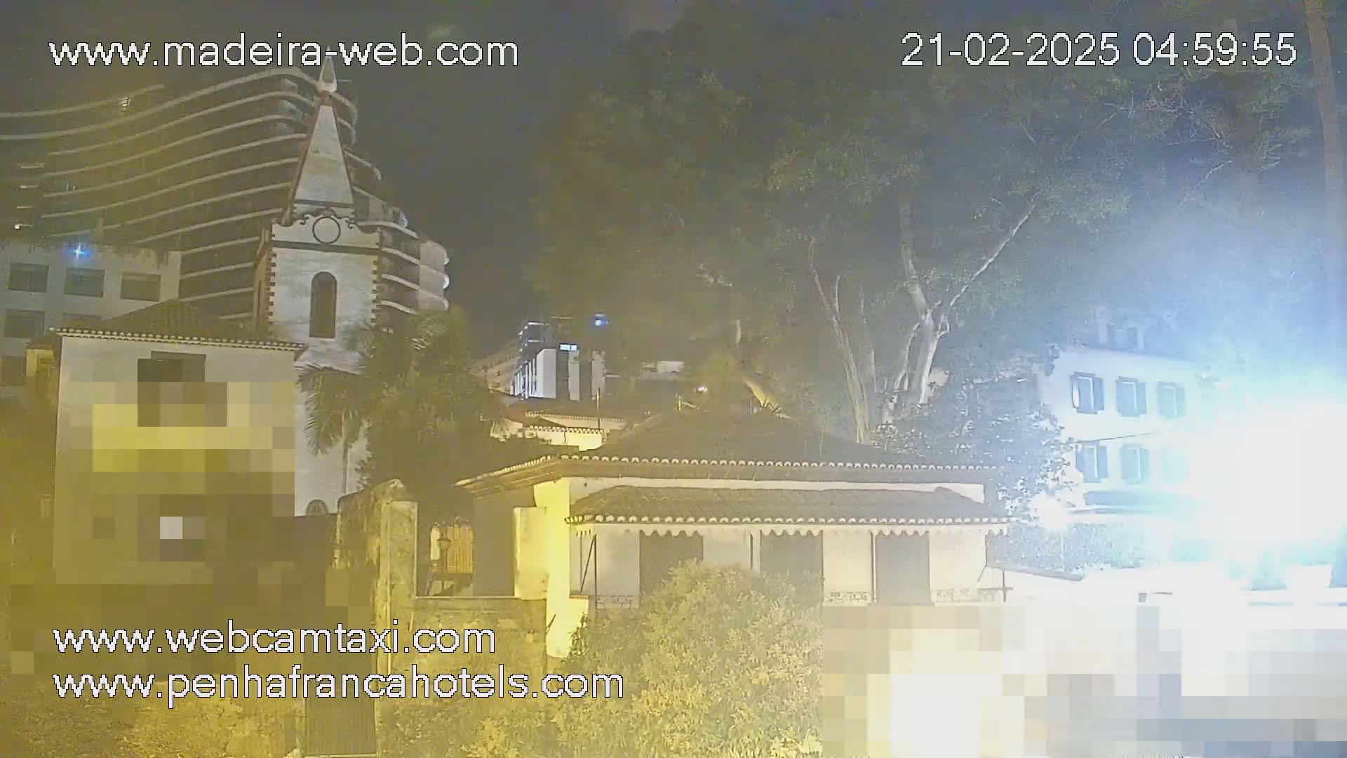 A night scene shows several buildings, including a tall, modern building and a smaller, older-looking building with a church-like tower, partially obscured by hazy, nighttime conditions and foliage.