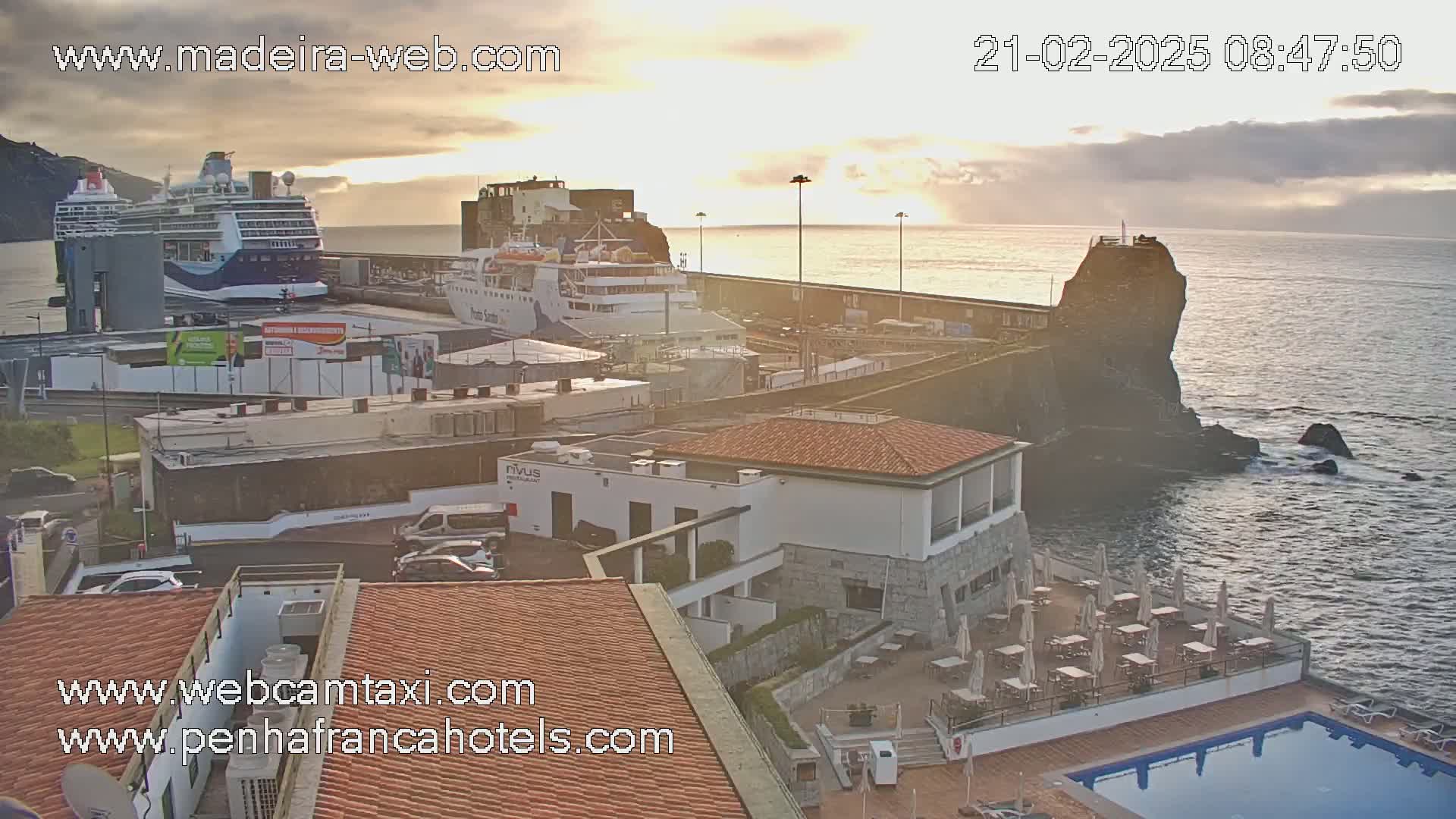 A harbor at sunrise, featuring two cruise ships docked, a rocky outcropping, and a building with an outdoor patio and pool.