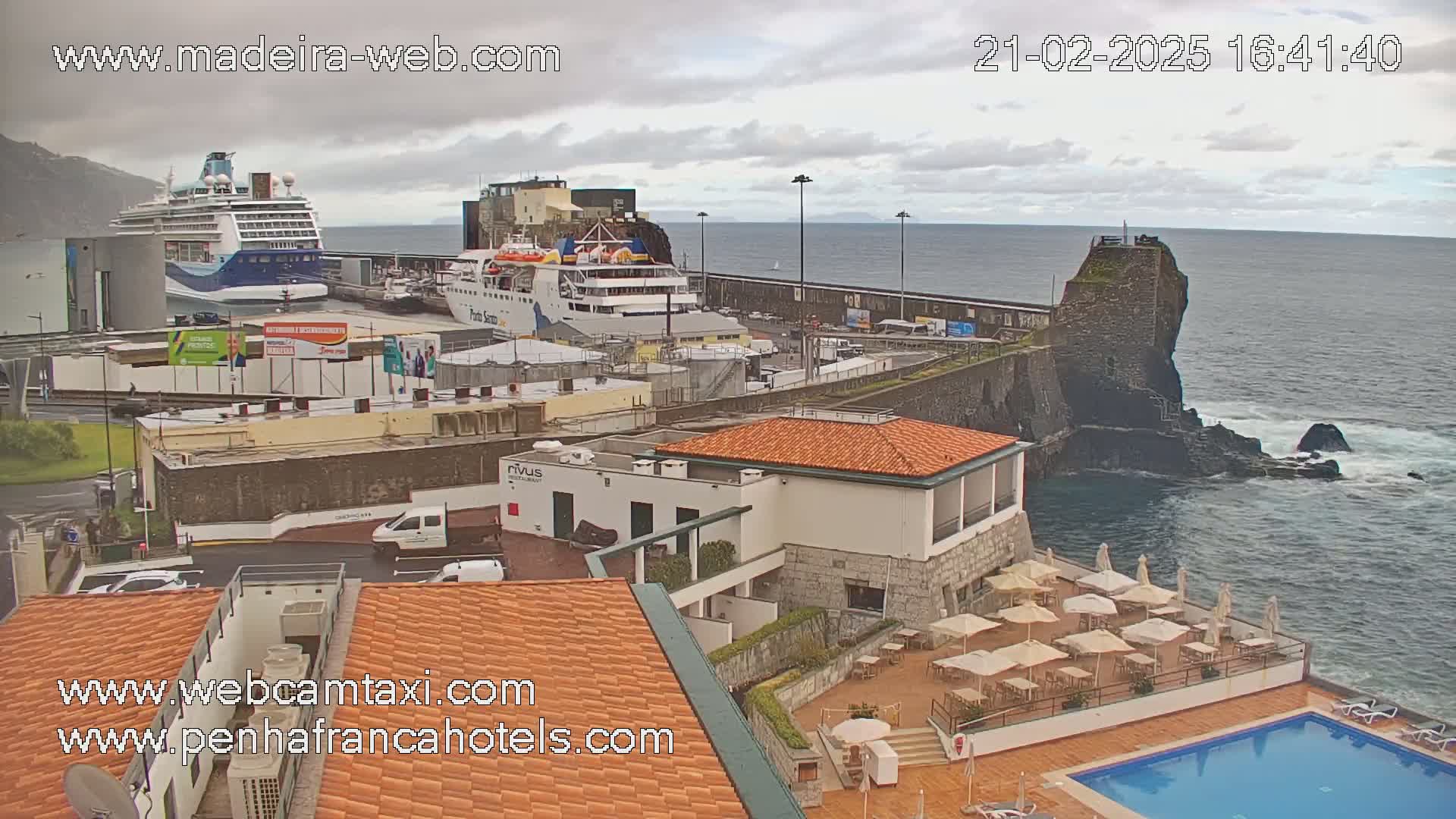 An overcast day at a harbor shows two large ships docked, buildings, a pool, and an outdoor patio area beside a rocky coastline.