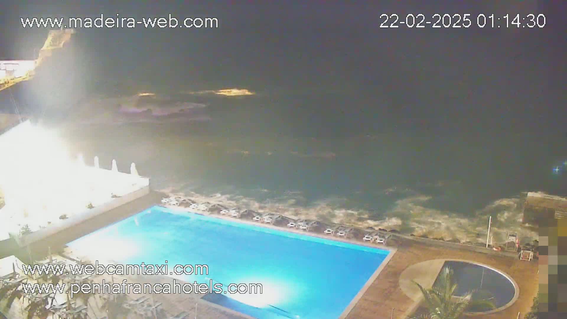 A nighttime view shows a swimming pool next to a turbulent ocean under a dark sky.