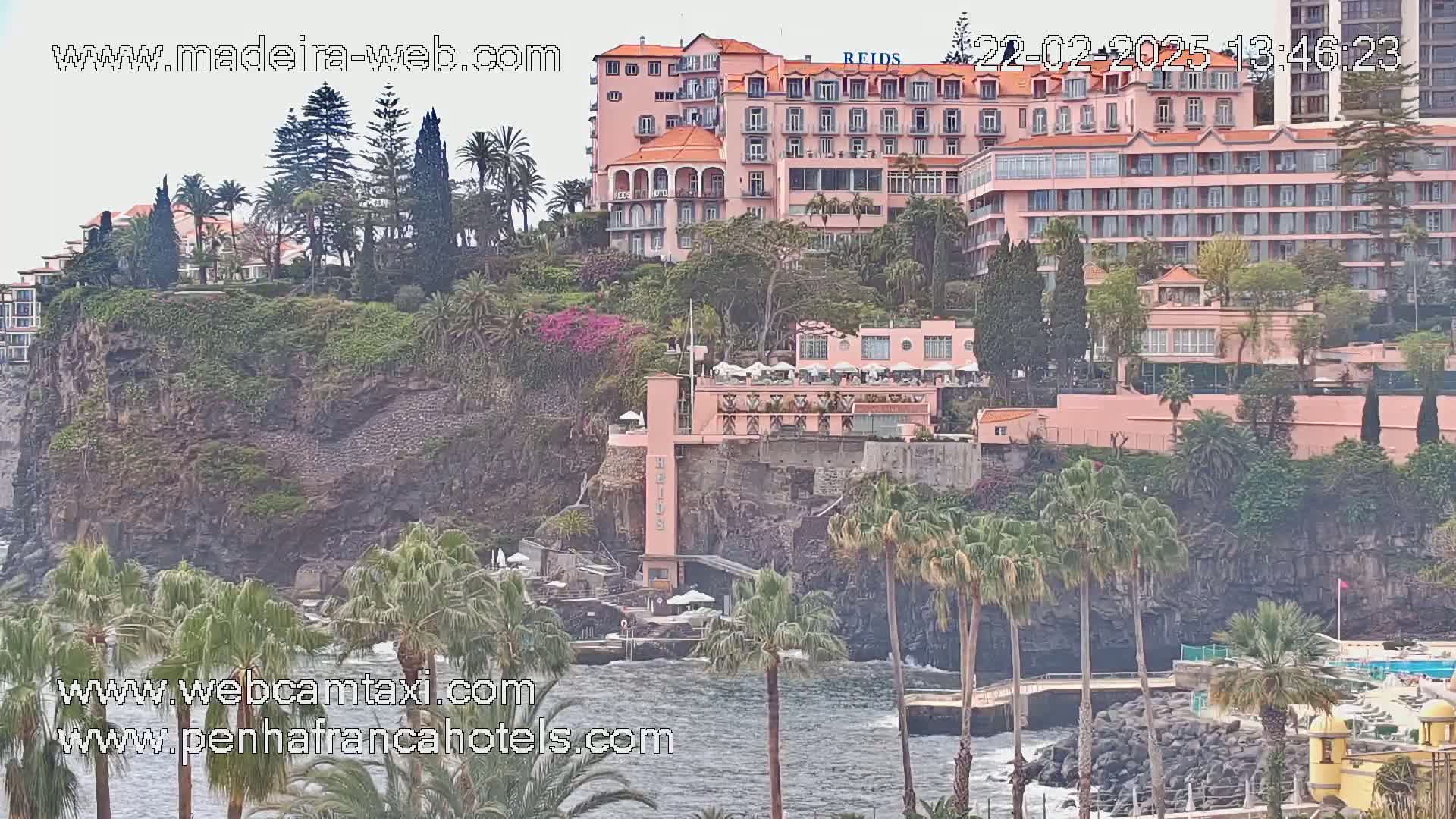 A pink hotel complex sits atop a rocky cliff overlooking a calm ocean dotted with palm trees under a mostly clear sky.