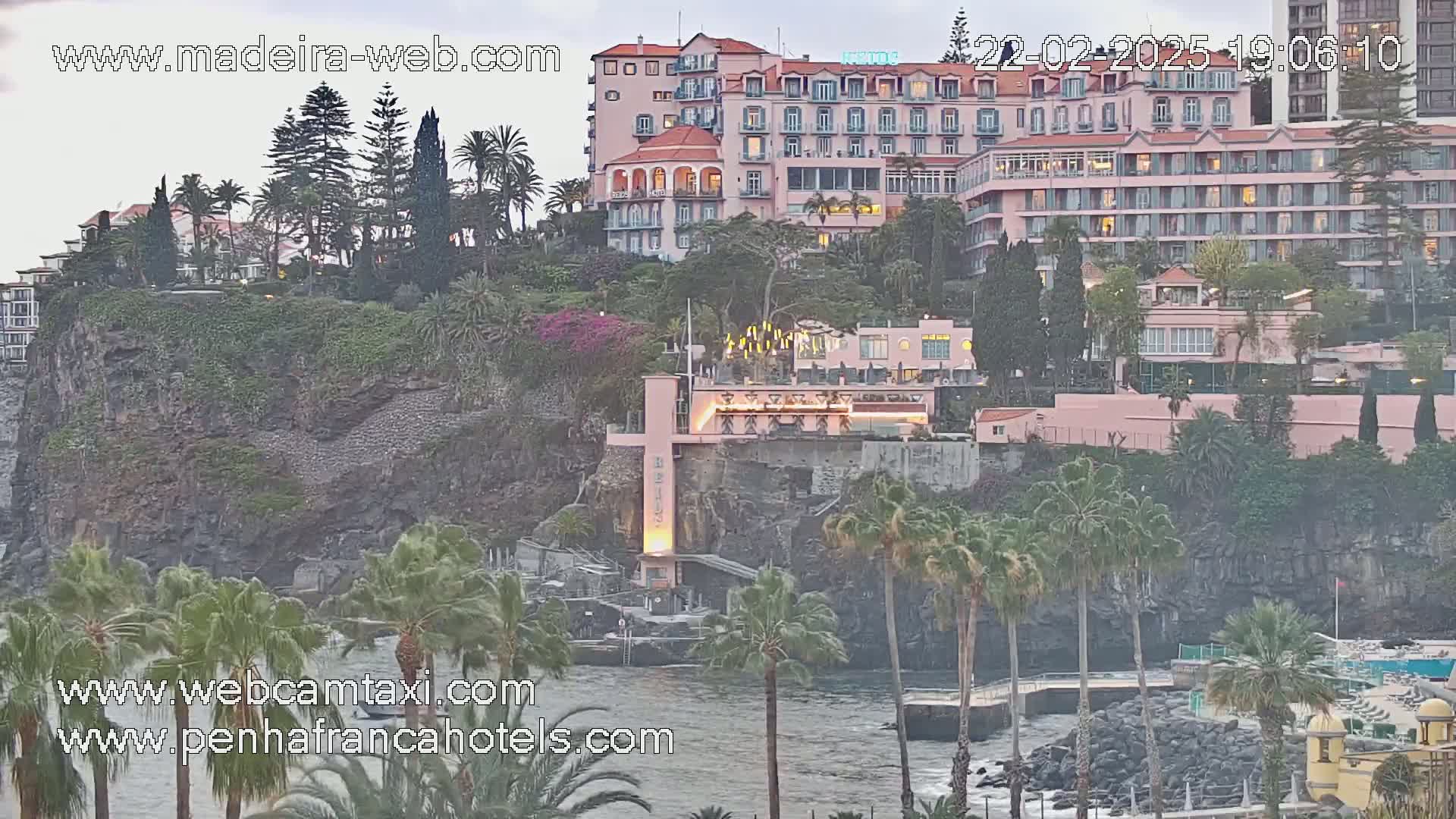 A cliffside hotel complex overlooks a rocky coastline at dusk, with palm trees dotting the landscape.