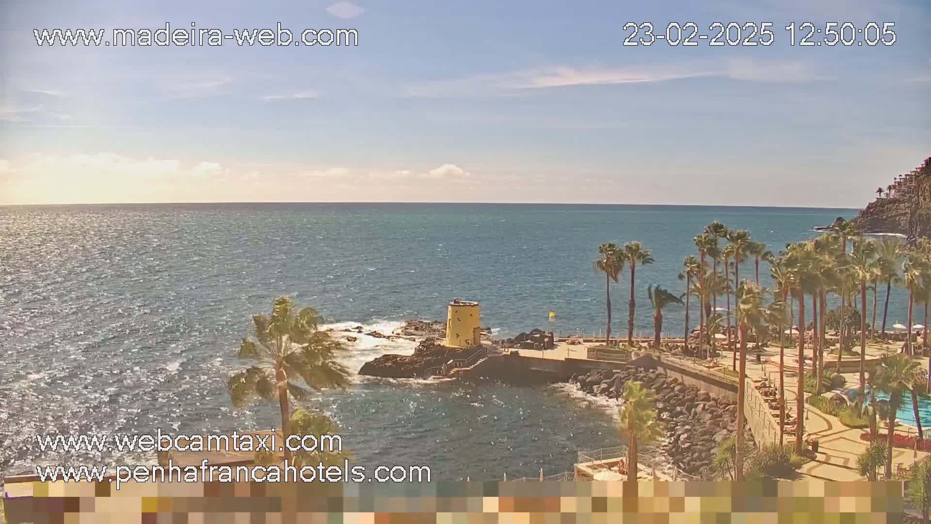 A partly sunny day shows a calm ocean with a rocky shoreline featuring palm trees, a small yellow tower, and a resort-like area.