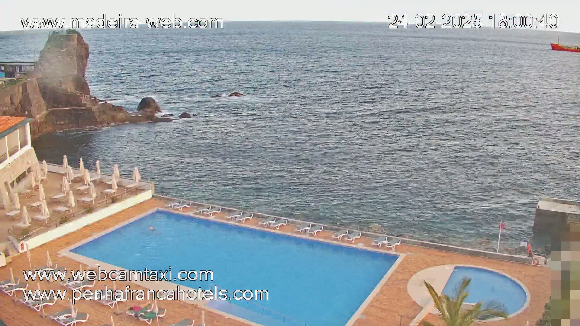An outdoor swimming pool and sunbathing area are situated next to a rocky coastline and ocean under a partly cloudy sky.