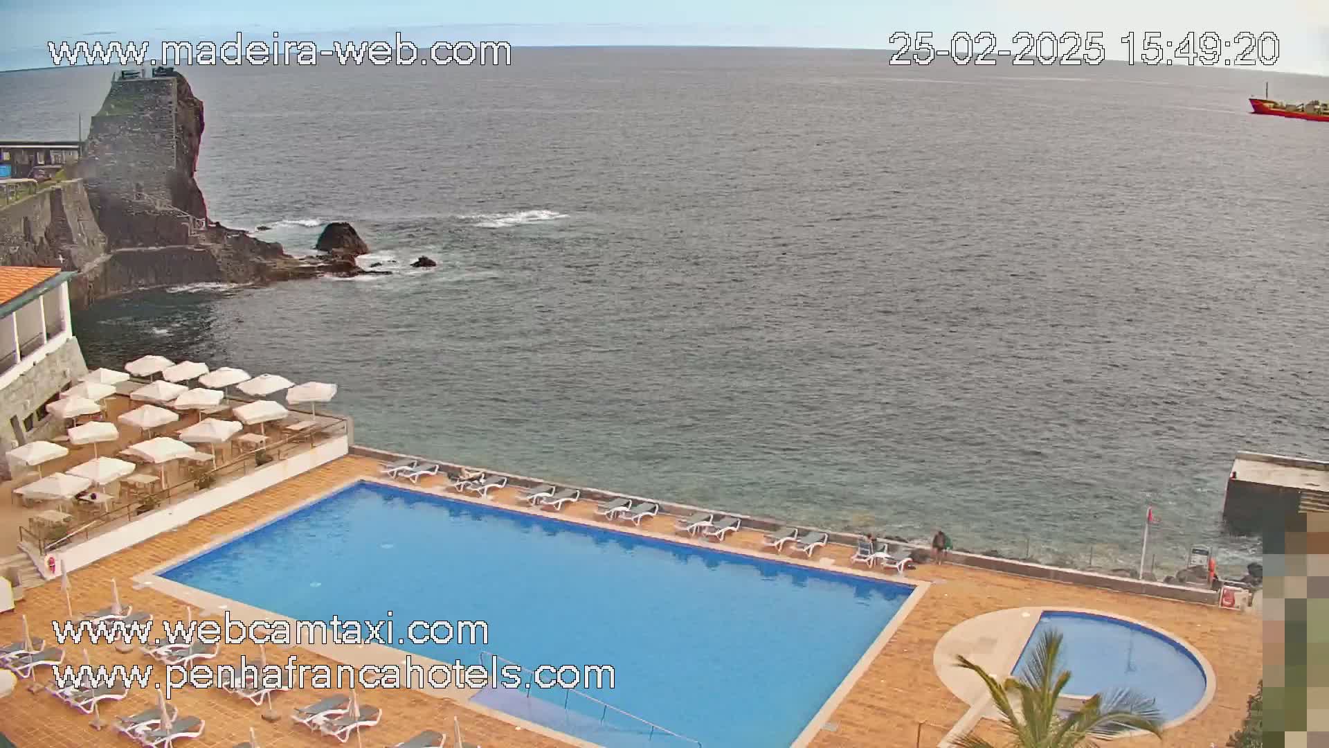 A hotel pool area with numerous umbrellas and lounge chairs overlooks a calm ocean and a rocky shoreline.