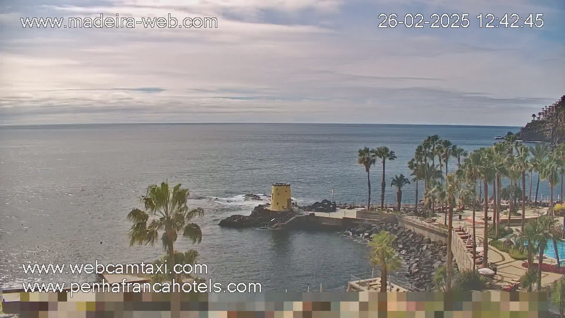 A partly cloudy day overlooks a calm ocean with a rocky shoreline lined with palm trees and a small yellow structure.