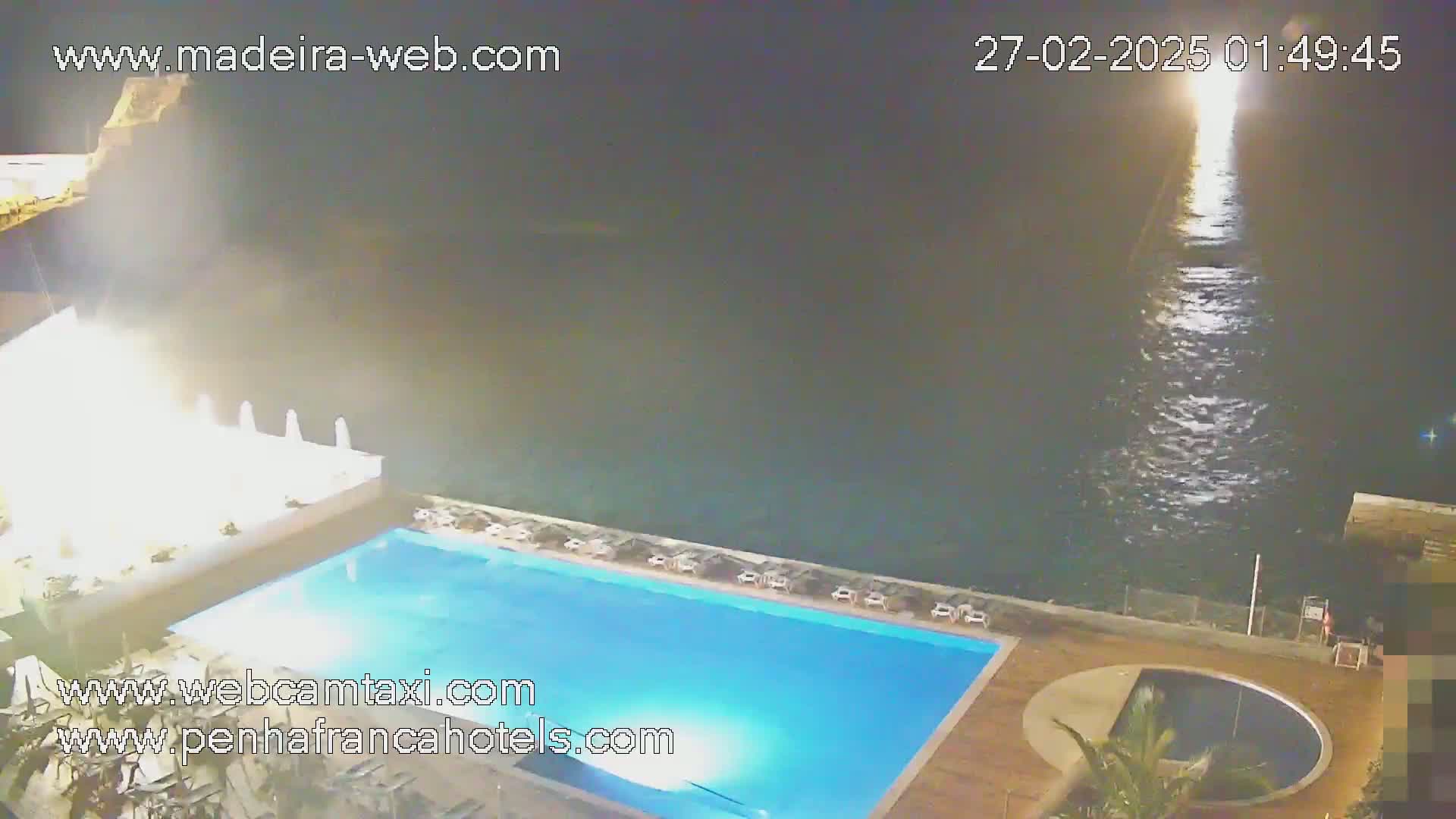 A nighttime view of a resort pool area overlooking a calm ocean under a dark sky.