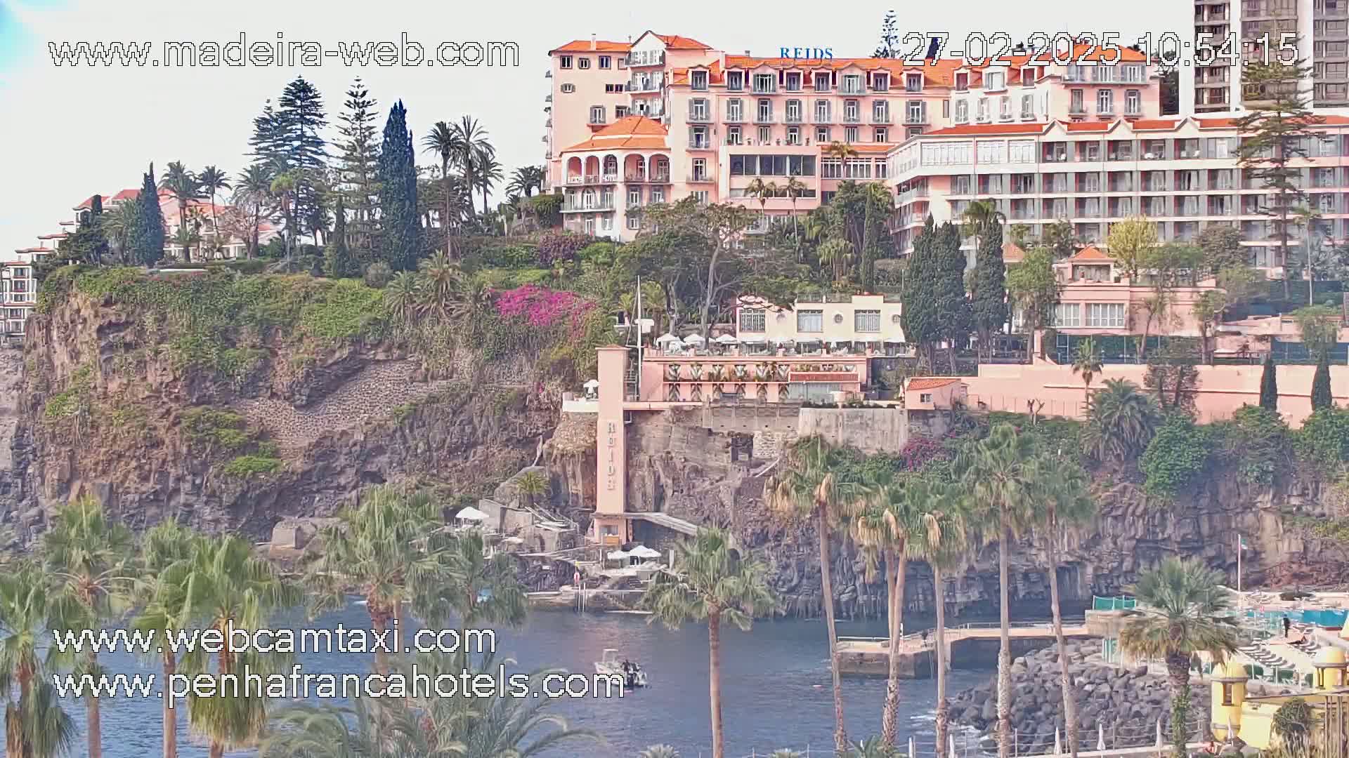 A coastal scene on a partly sunny day shows large hotels and buildings on a cliff overlooking a calm body of water with palm trees and lush vegetation.