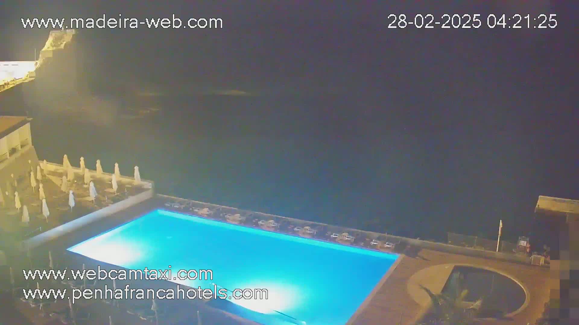 A nighttime aerial view shows a lit swimming pool and surrounding patio area next to a dark ocean.