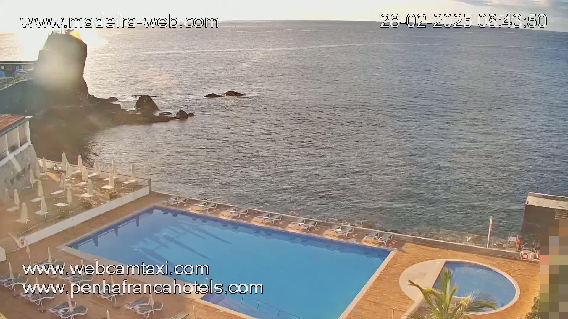 A hotel pool and patio area overlook a calm ocean with a rocky outcropping in the distance under a mostly clear sky.
