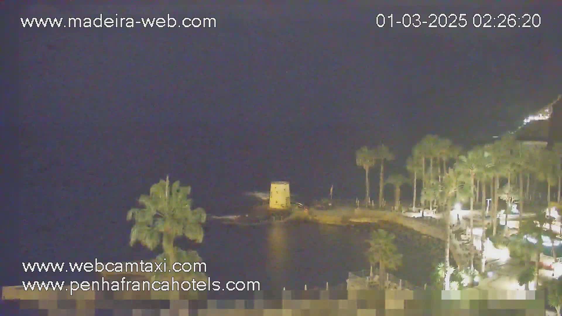 A nighttime coastal scene shows a small, illuminated structure on a dark body of water, surrounded by palm trees and a dimly lit walkway.