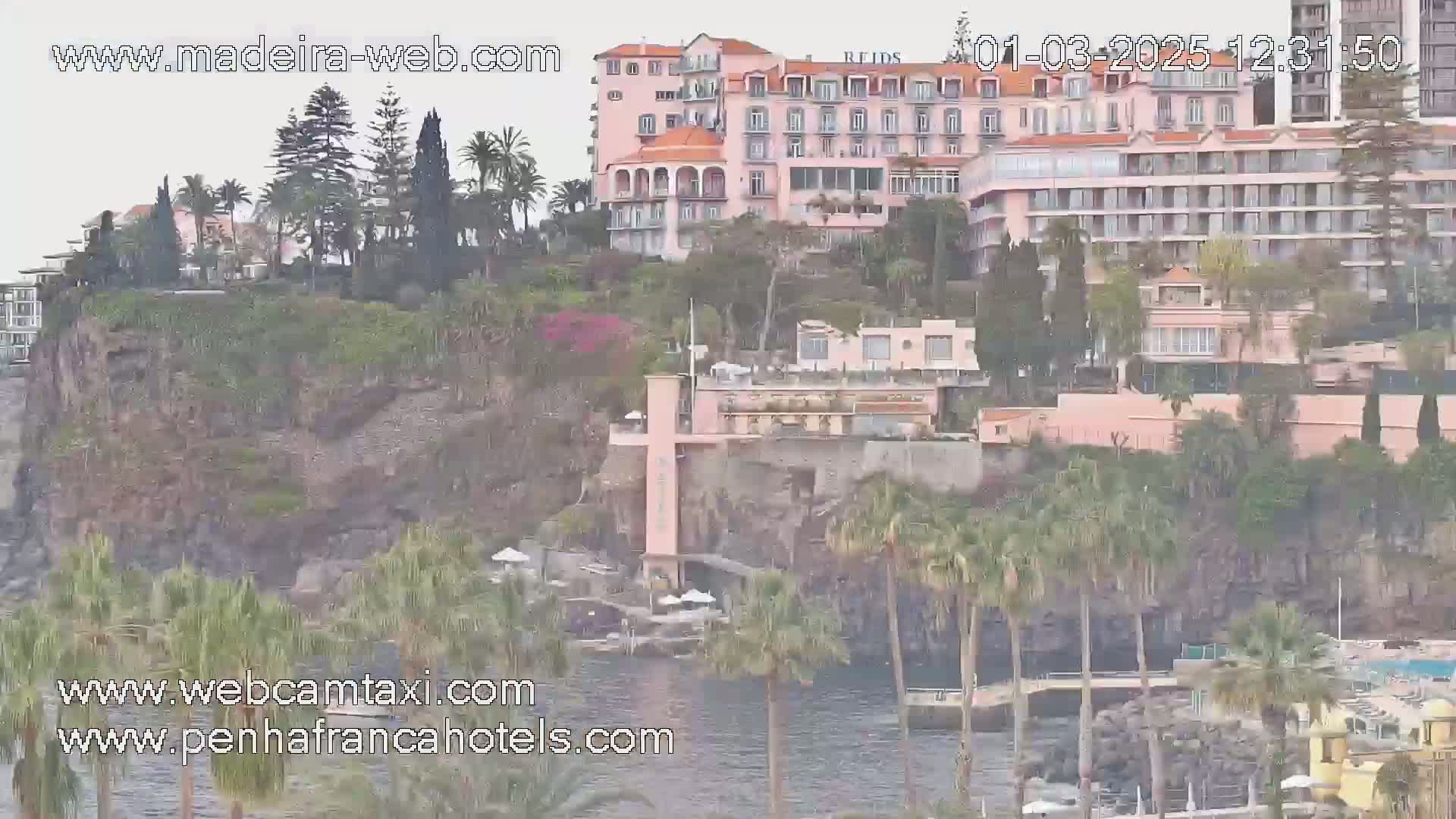A large pink hotel complex is situated on a cliff overlooking a calm body of water under an overcast sky.