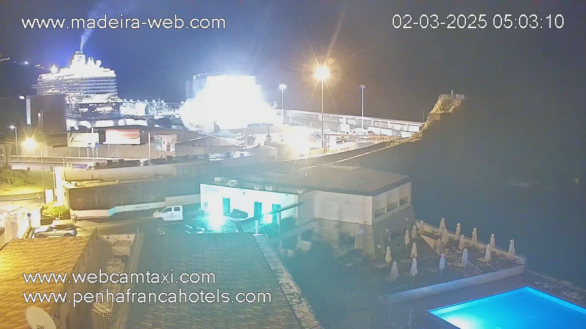 A large cruise ship is docked at a port at night, with a hotel featuring a pool and outdoor umbrellas visible in the foreground.