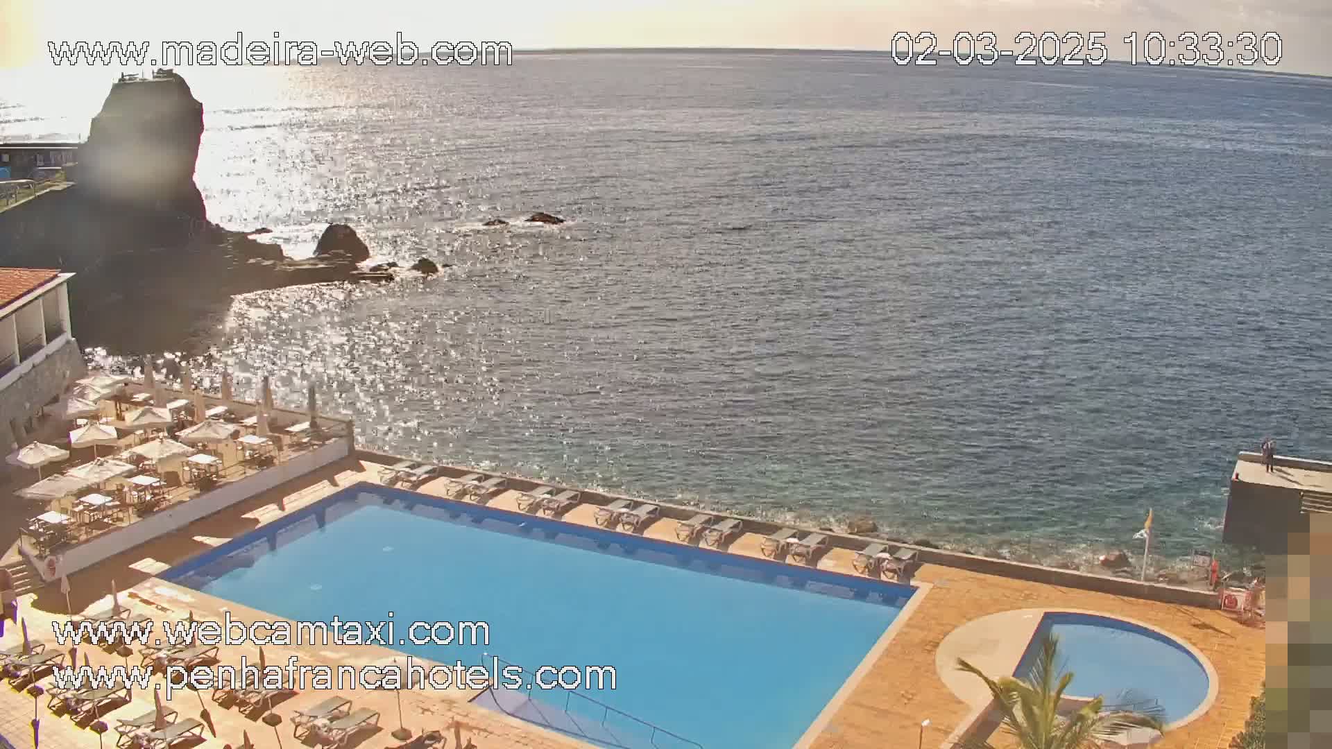 A resort pool area with sun loungers and a restaurant overlooking a calm ocean on a sunny day.