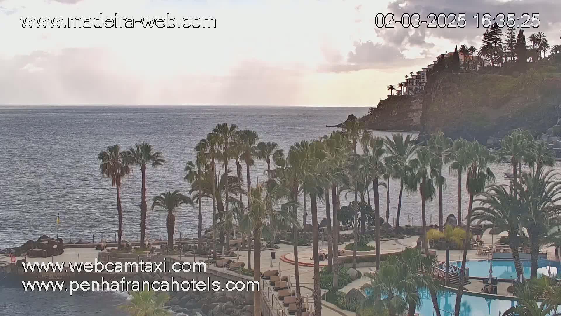 A resort with palm trees and swimming pools overlooks a calm ocean under a partly cloudy sky.
