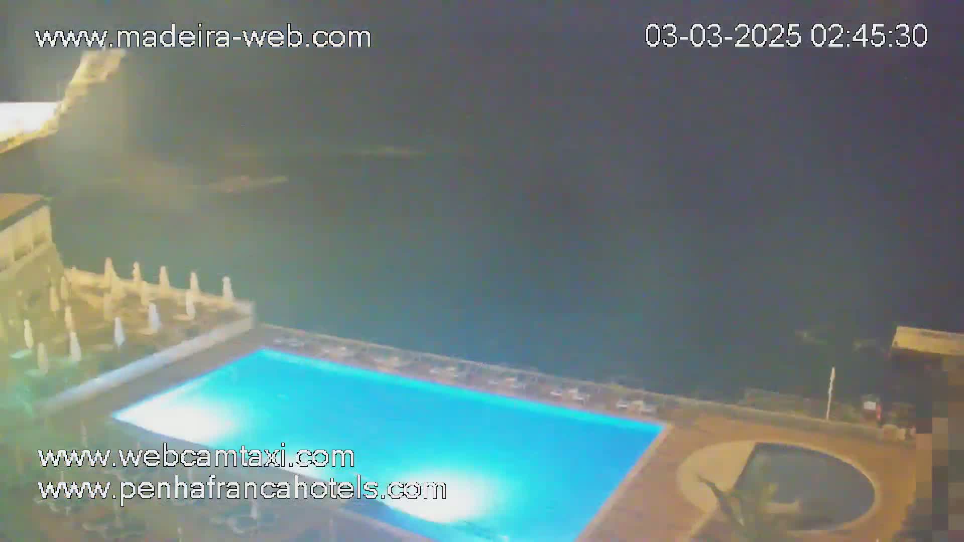 An outdoor swimming pool at night, illuminated, with closed parasols surrounding it and a dark ocean visible in the background.