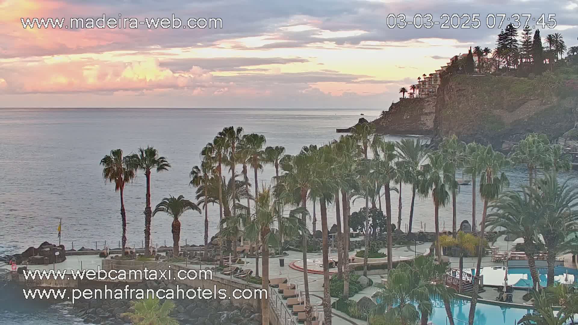A coastal resort with palm trees and pools is seen at sunrise under a partly cloudy sky.