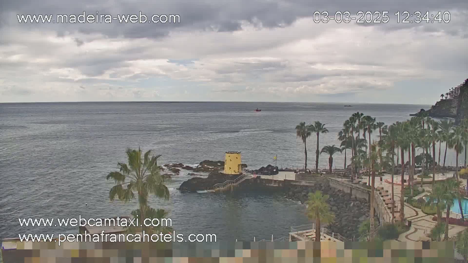 A partly cloudy day shows a rocky coast with palm trees, a small yellow tower, and a calm ocean with a distant vessel.