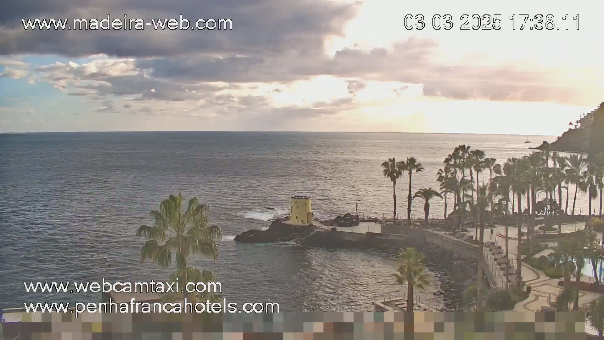 A partly cloudy sunset over a calm ocean with palm trees lining a rocky shoreline and a small yellow structure on a point of land.