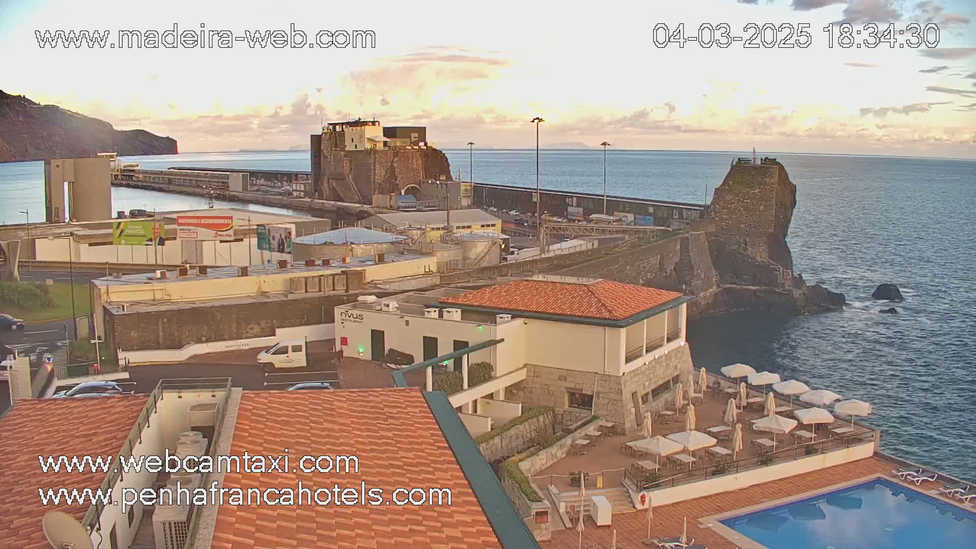 A coastal town at sunset with calm waters, featuring a harbor, buildings, a swimming pool, and patio furniture.