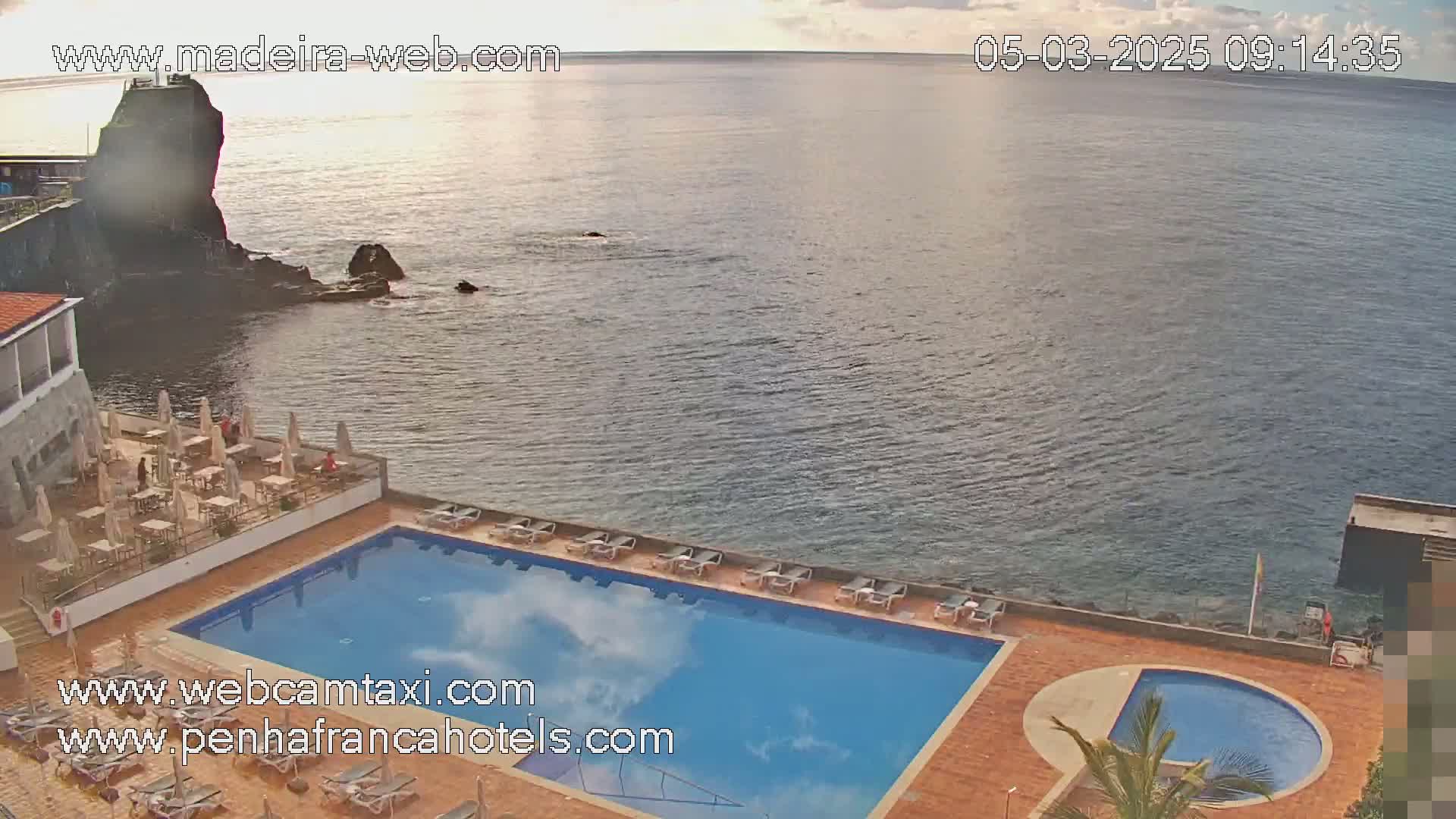 An outdoor swimming pool and smaller circular pool are situated on a hotel patio overlooking a calm ocean under a partly cloudy sky.
