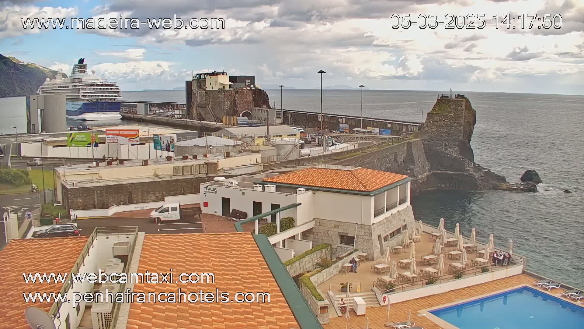 A cruise ship is docked at a port city next to a rocky coastline under a partly cloudy sky.