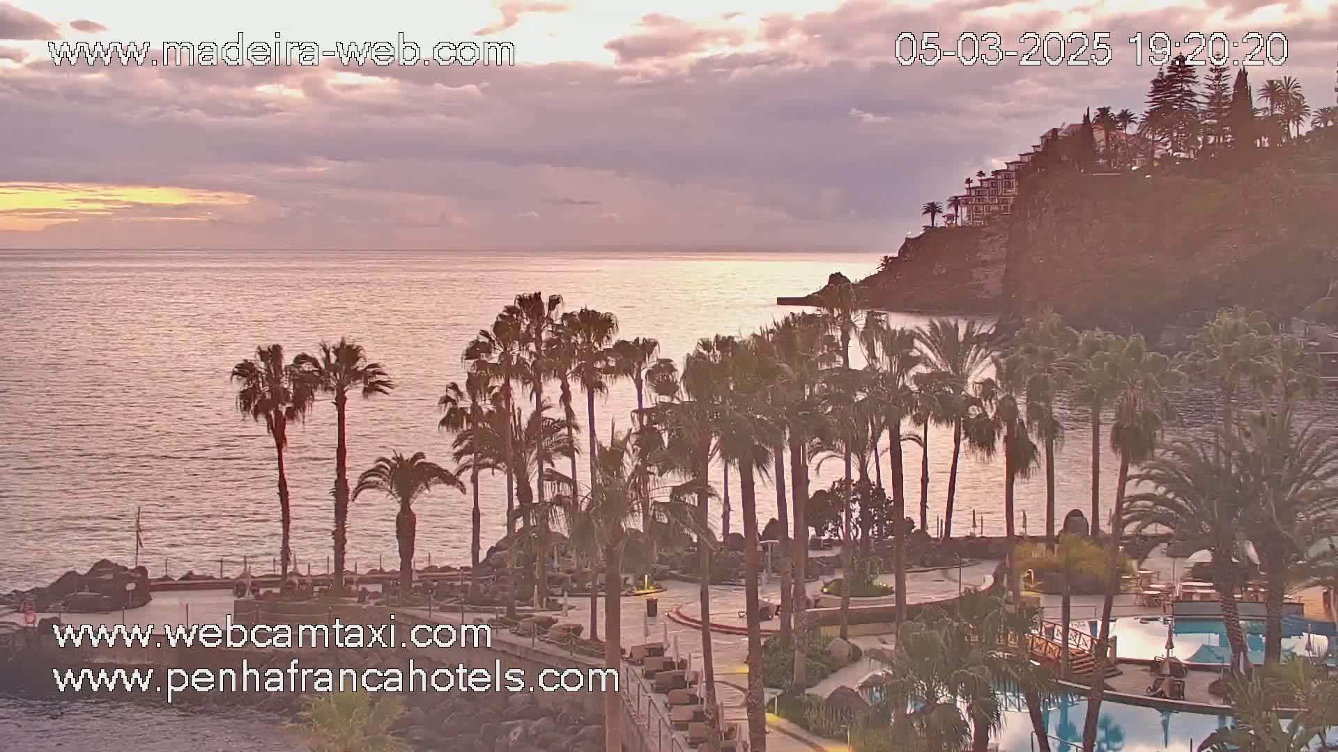 A calm ocean at sunset is seen from a seaside resort with palm trees and swimming pools, under a partly cloudy sky.