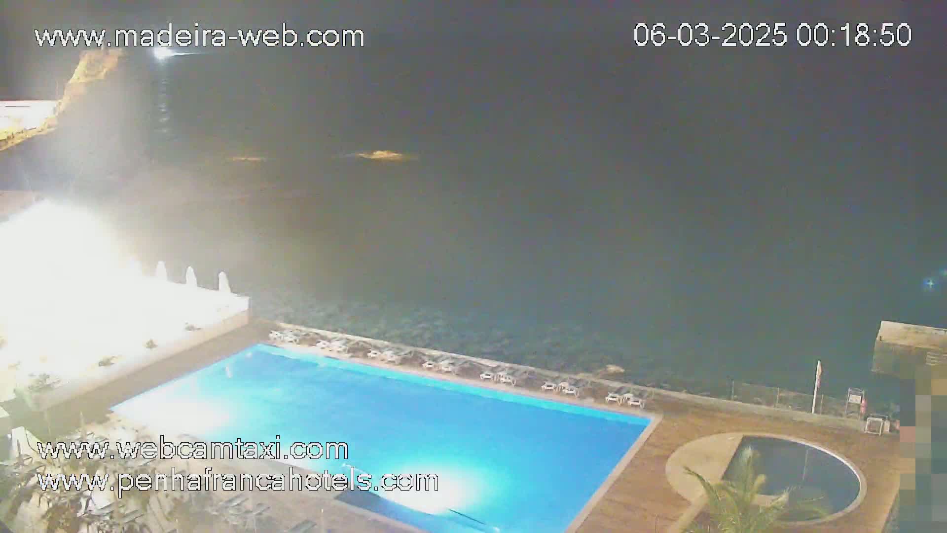 An illuminated swimming pool and a smaller, round pool are situated on a deck overlooking a dark ocean at night.