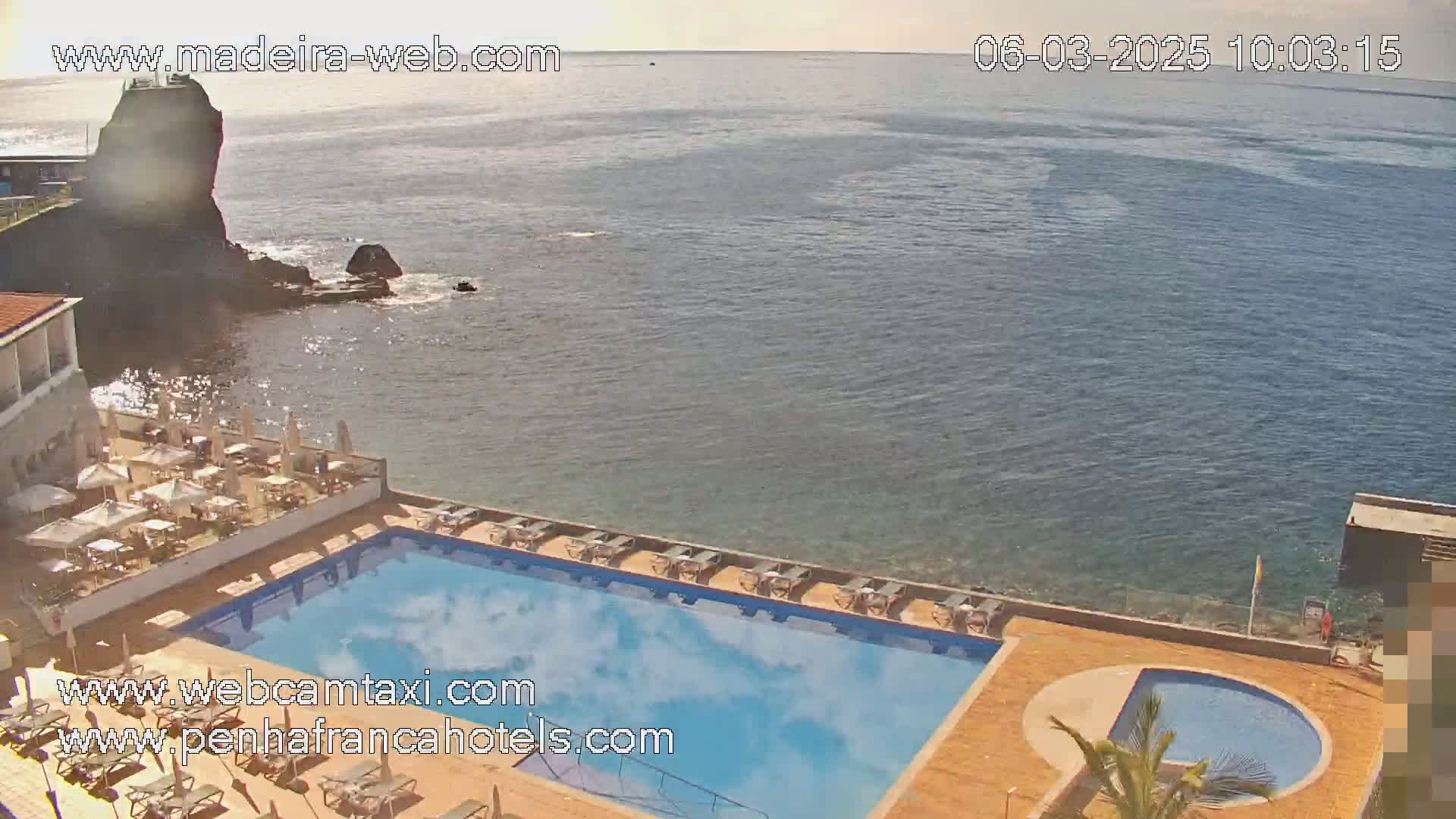 A swimming pool and patio area overlook a calm ocean on a sunny day, with a rocky outcropping visible in the distance.