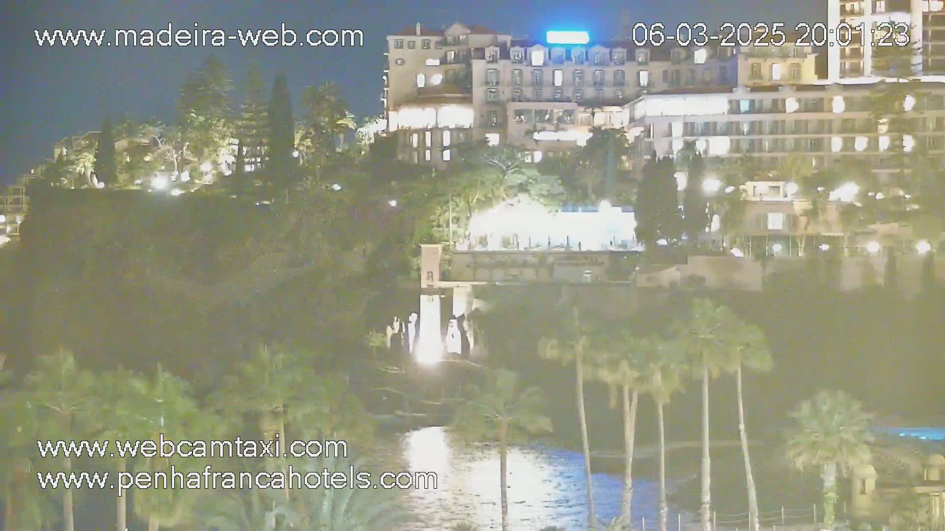 A nighttime view of a brightly lit hotel situated on a hill overlooking a body of water, surrounded by palm trees and lush vegetation.