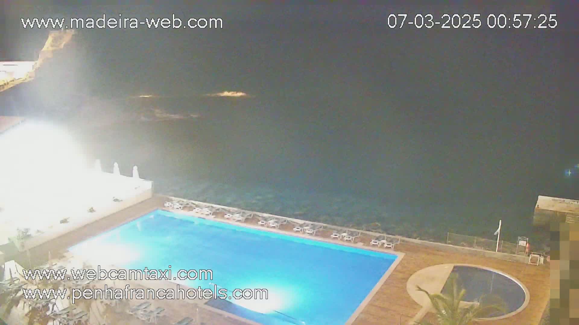 An outdoor swimming pool and a smaller round pool are illuminated at night next to a dark ocean.