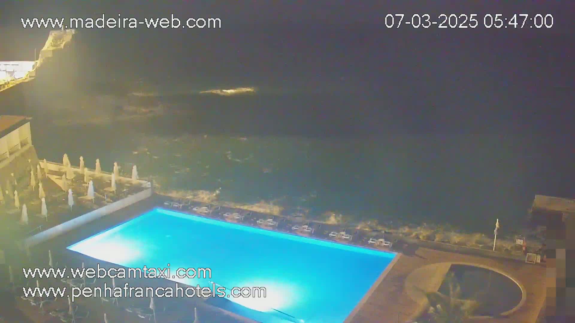 An illuminated swimming pool sits beside a dark ocean with waves crashing against the shore at night.