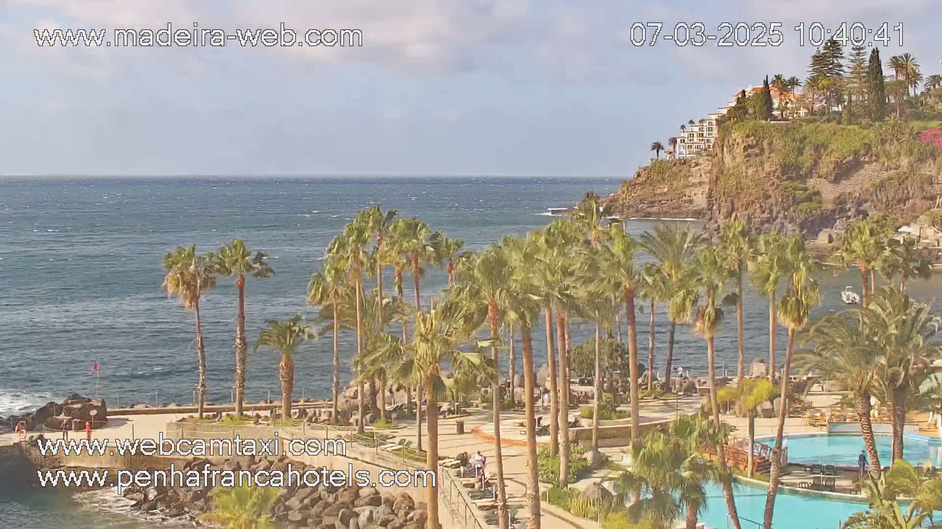 A tropical resort with palm trees and swimming pools overlooks a calm ocean under a partly cloudy sky.
