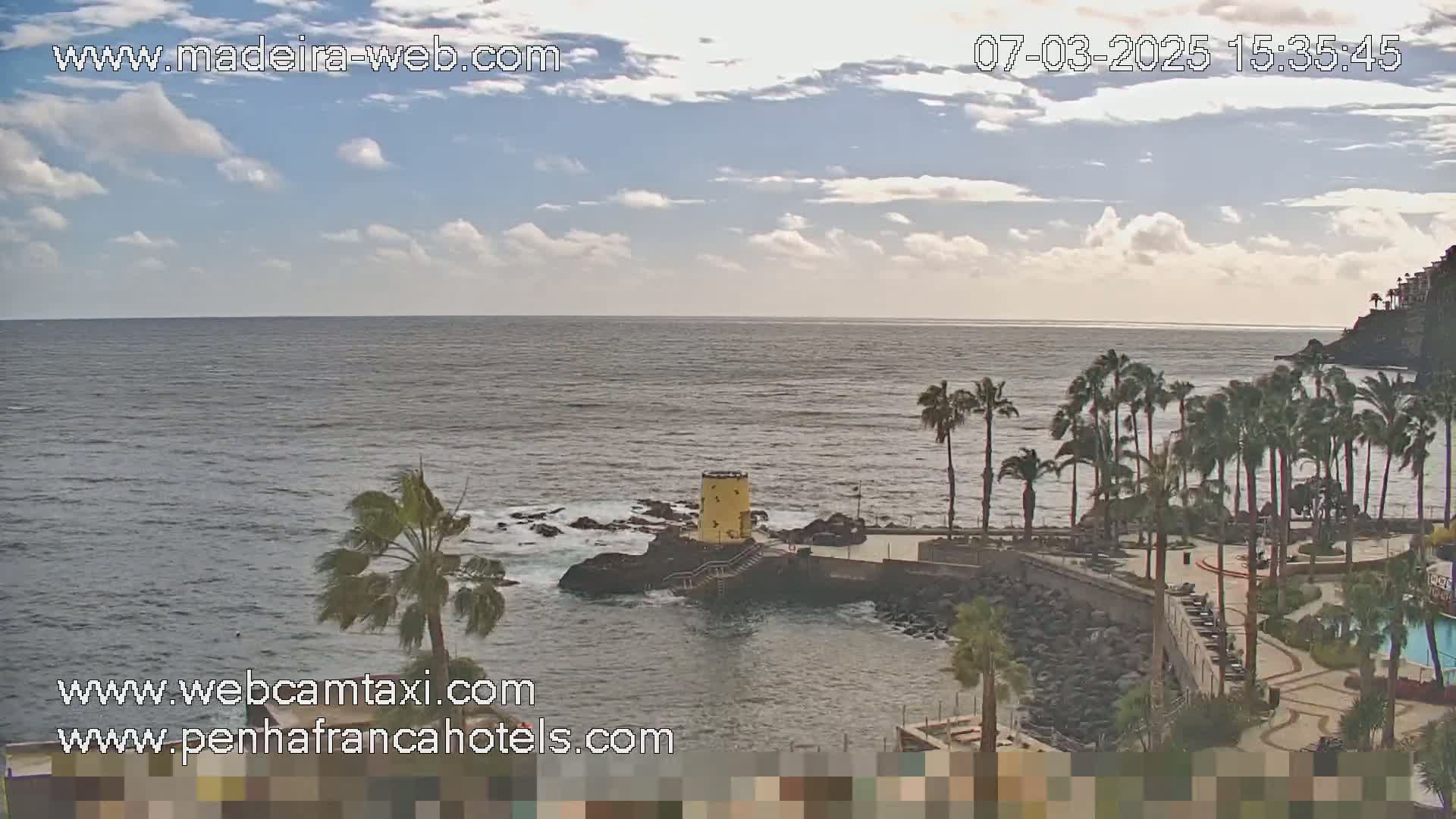 A partly cloudy day shows a rocky coast with palm trees, a small yellow tower, and a calm ocean.