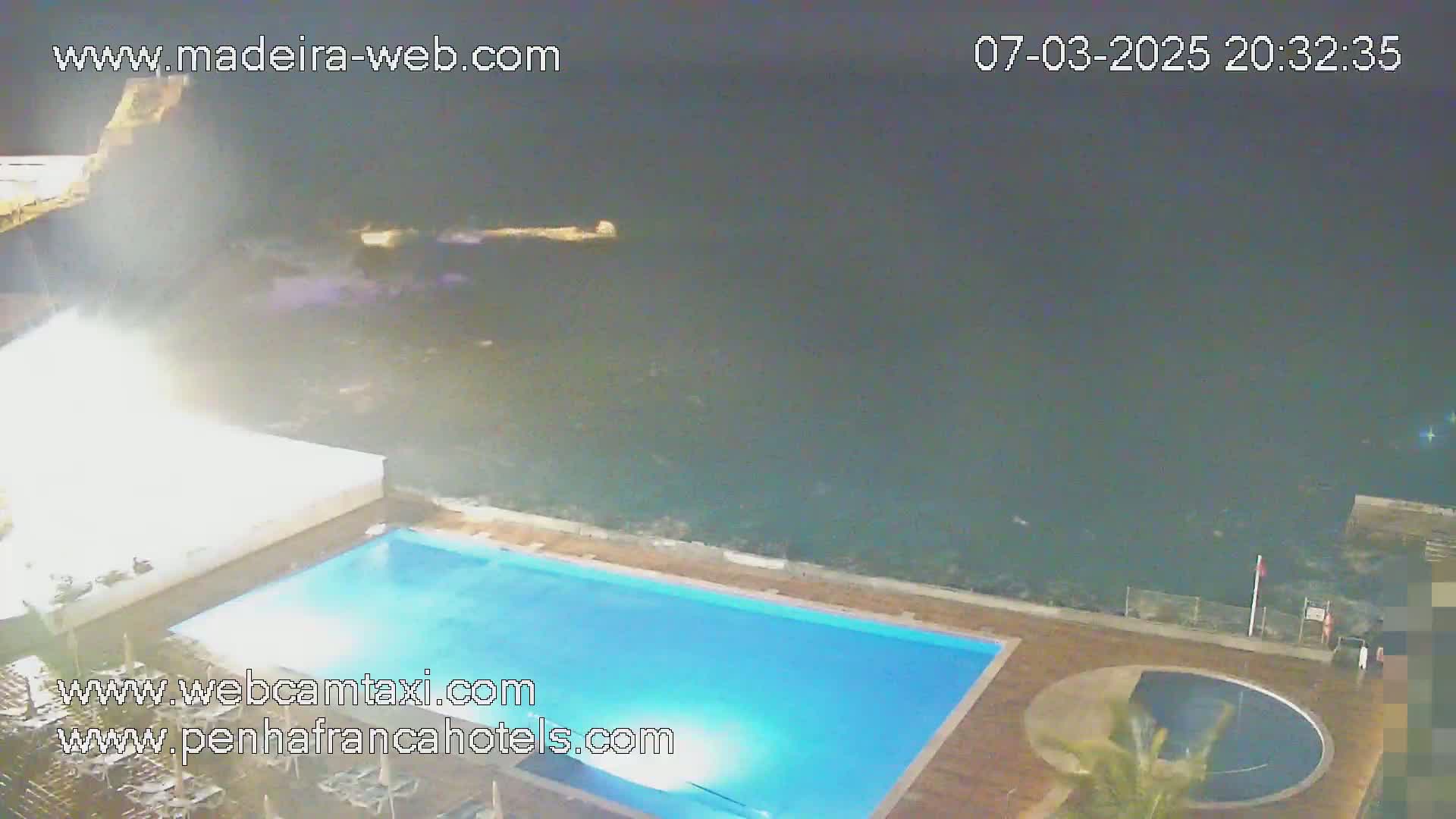 An illuminated swimming pool sits next to a dark ocean under a nighttime sky.